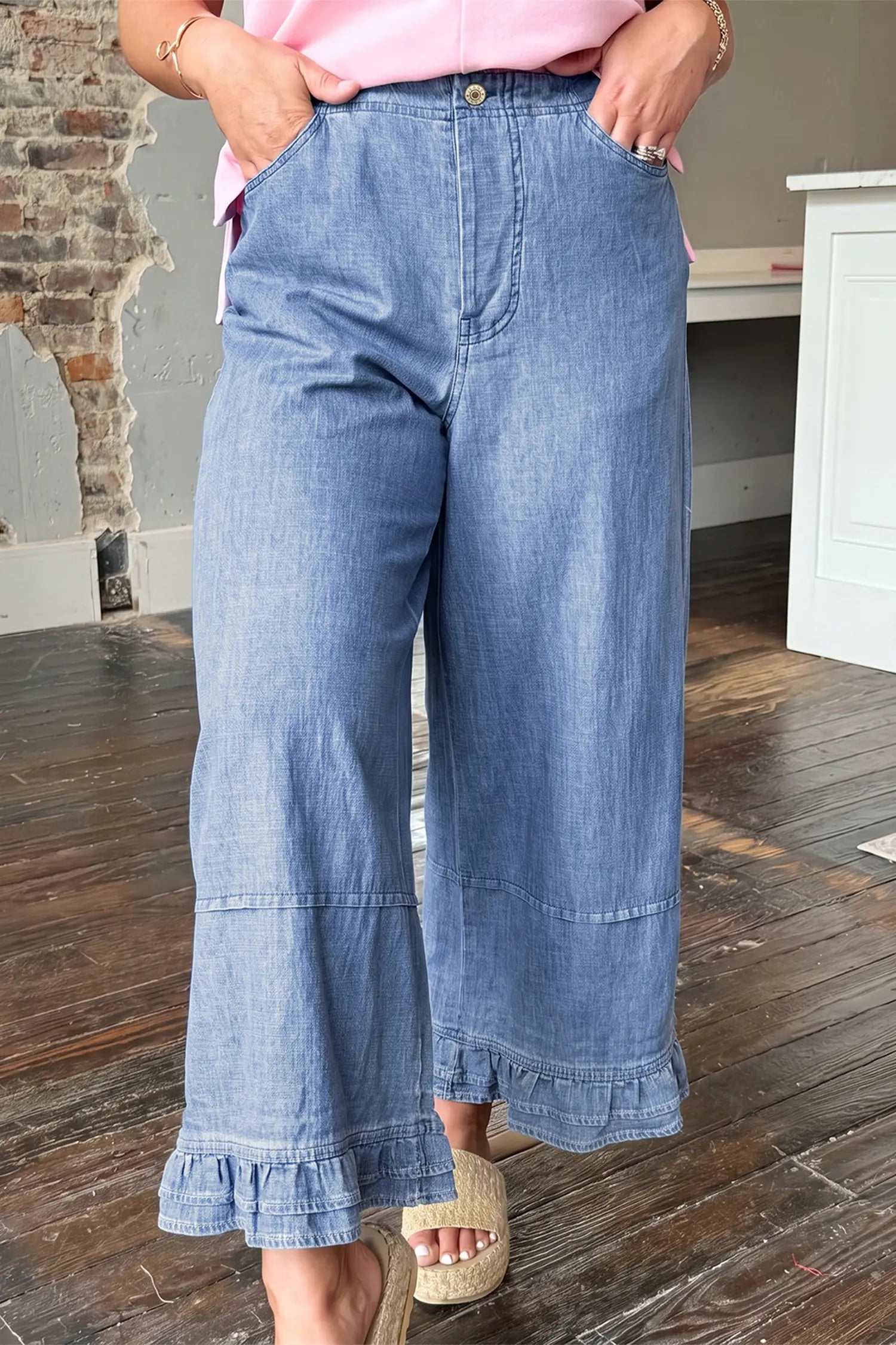 A person wearing light blue wide-leg denim pants with a ruffled hem, paired with a pink top and beige slide sandals, standing on a wooden floor in an indoor room.