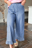 A person wearing light blue wide-leg denim pants with a ruffled hem, paired with a pink top and beige slide sandals, standing on a wooden floor in an indoor room.