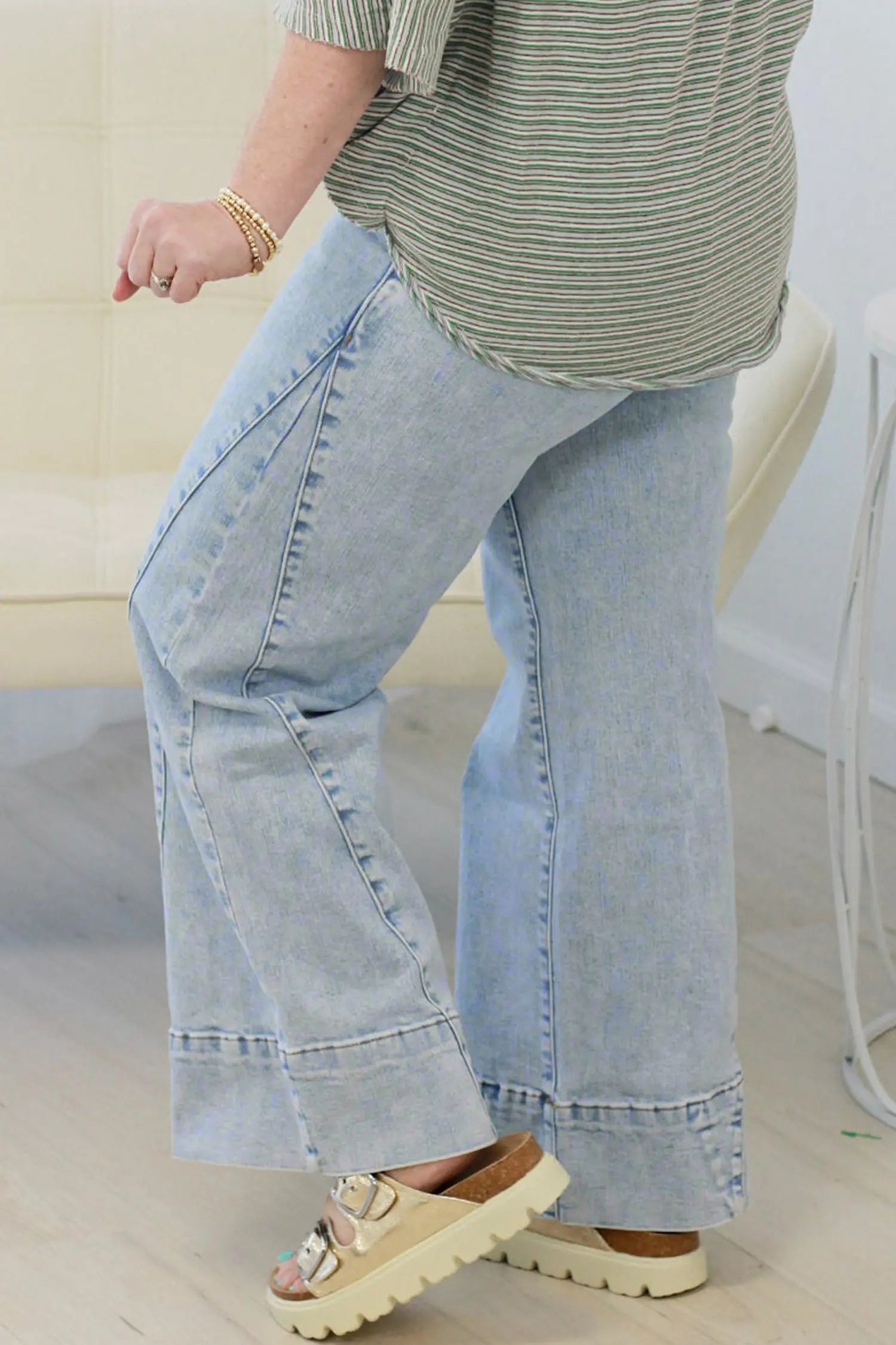 A person wearing light-wash wide-leg denim jeans with rolled cuffs, paired with a striped top and beige platform sandals, standing indoors.