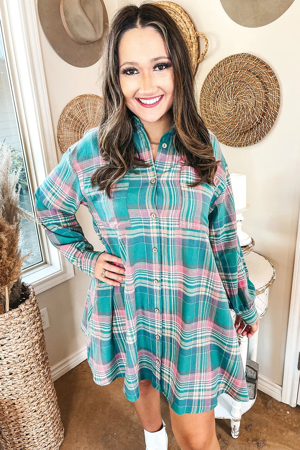 A woman wearing a teal, pink, and white plaid long-sleeve button-up shirt dress, standing in a decorated room with woven wall decor.