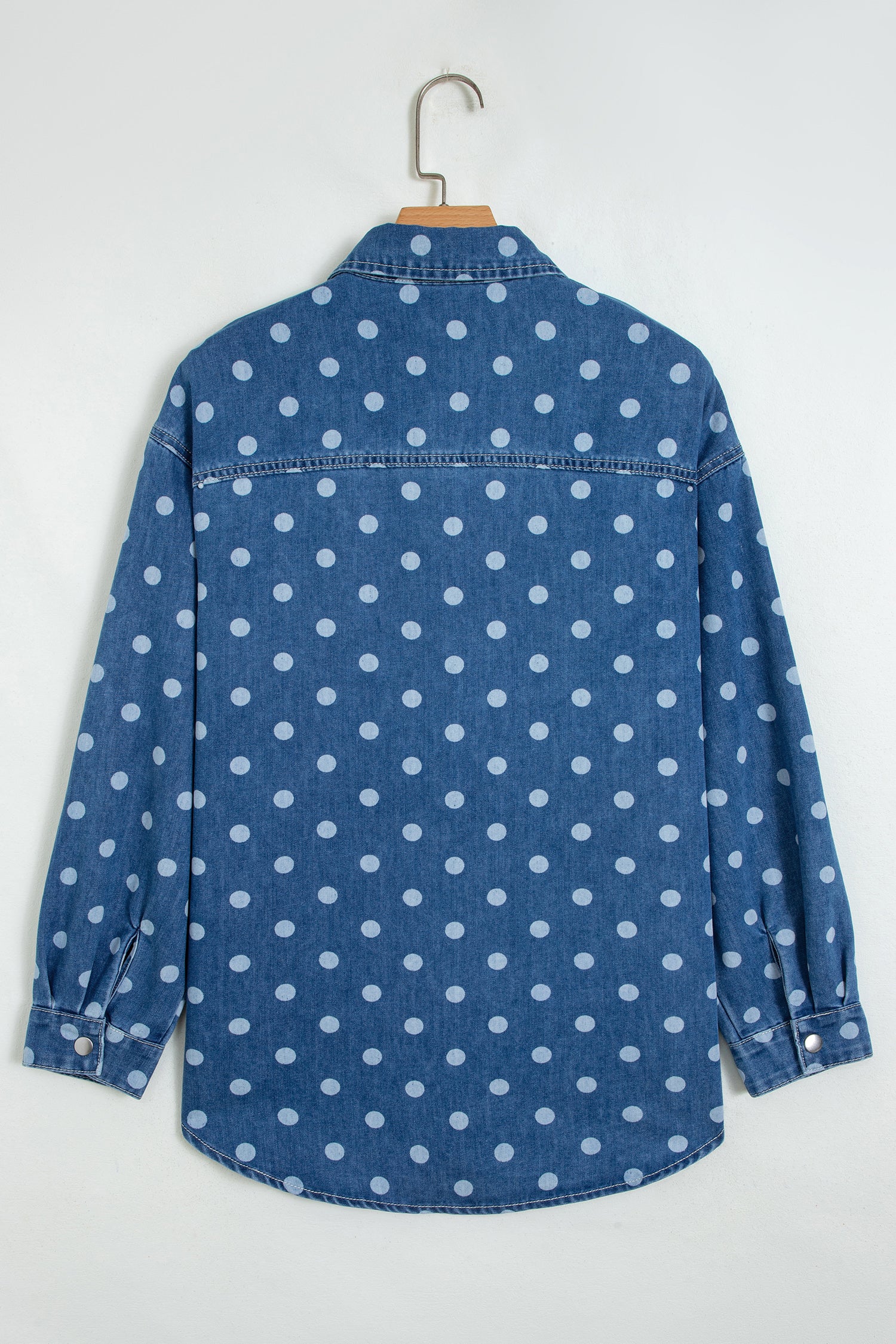 Blue denim long-sleeve shirt with white polka dots hanging on a wooden hanger against a white wall.