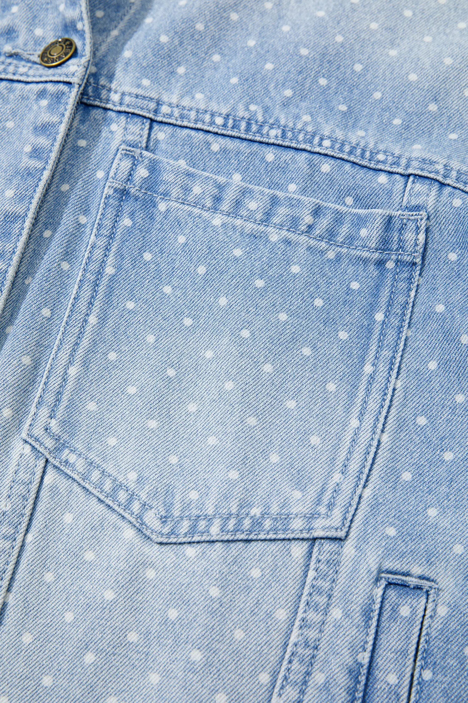 Close-up of a light blue denim jacket featuring a white polka dot pattern and a single front patch pocket.