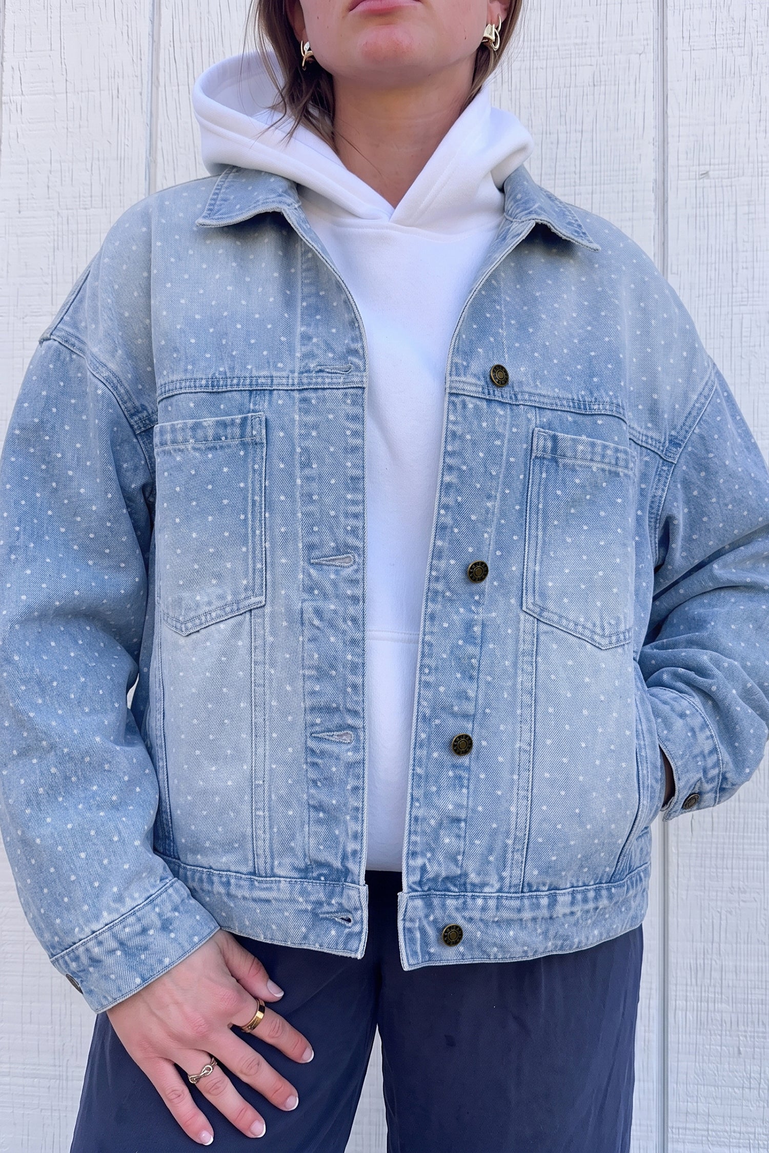 Person wearing light blue polka-dot denim jacket over white hoodie and navy pants against paneled wall.
