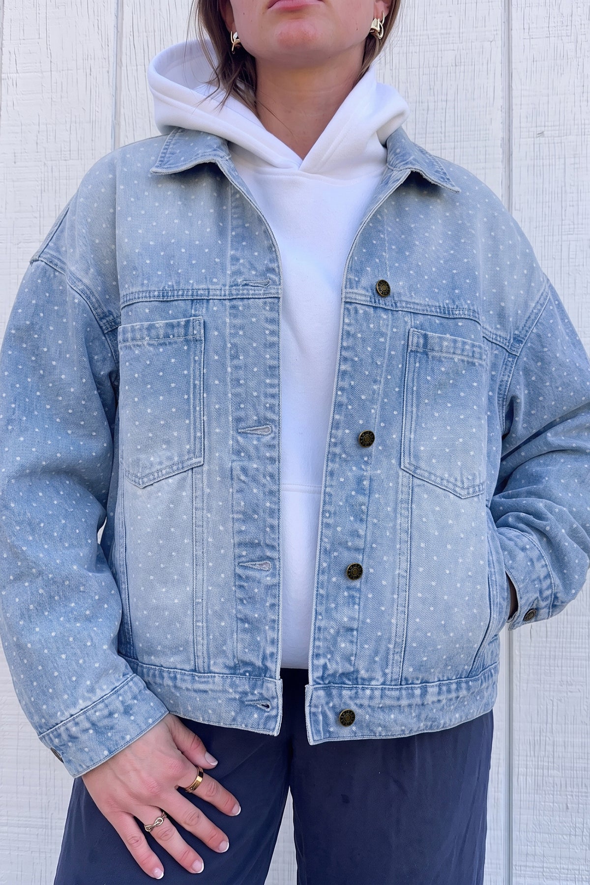 Person wearing light blue polka-dot denim jacket over white hoodie and navy pants against paneled wall.