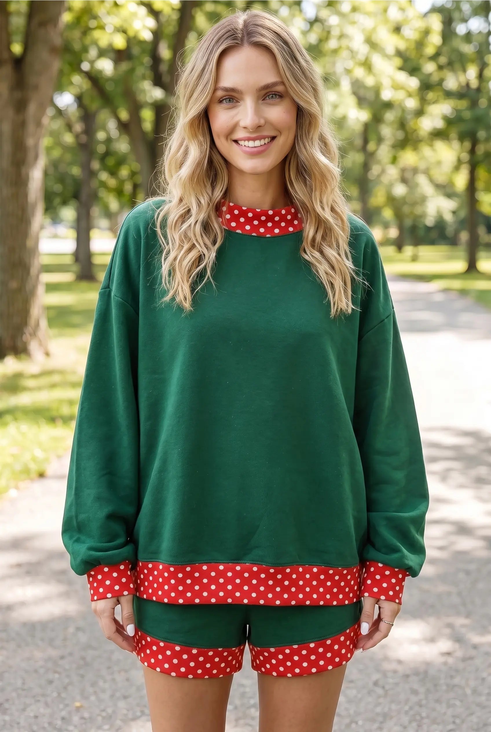 A woman wearing a dark green holiday-themed lounge set with red and white polka dot trim, standing on a paved path in a park.
