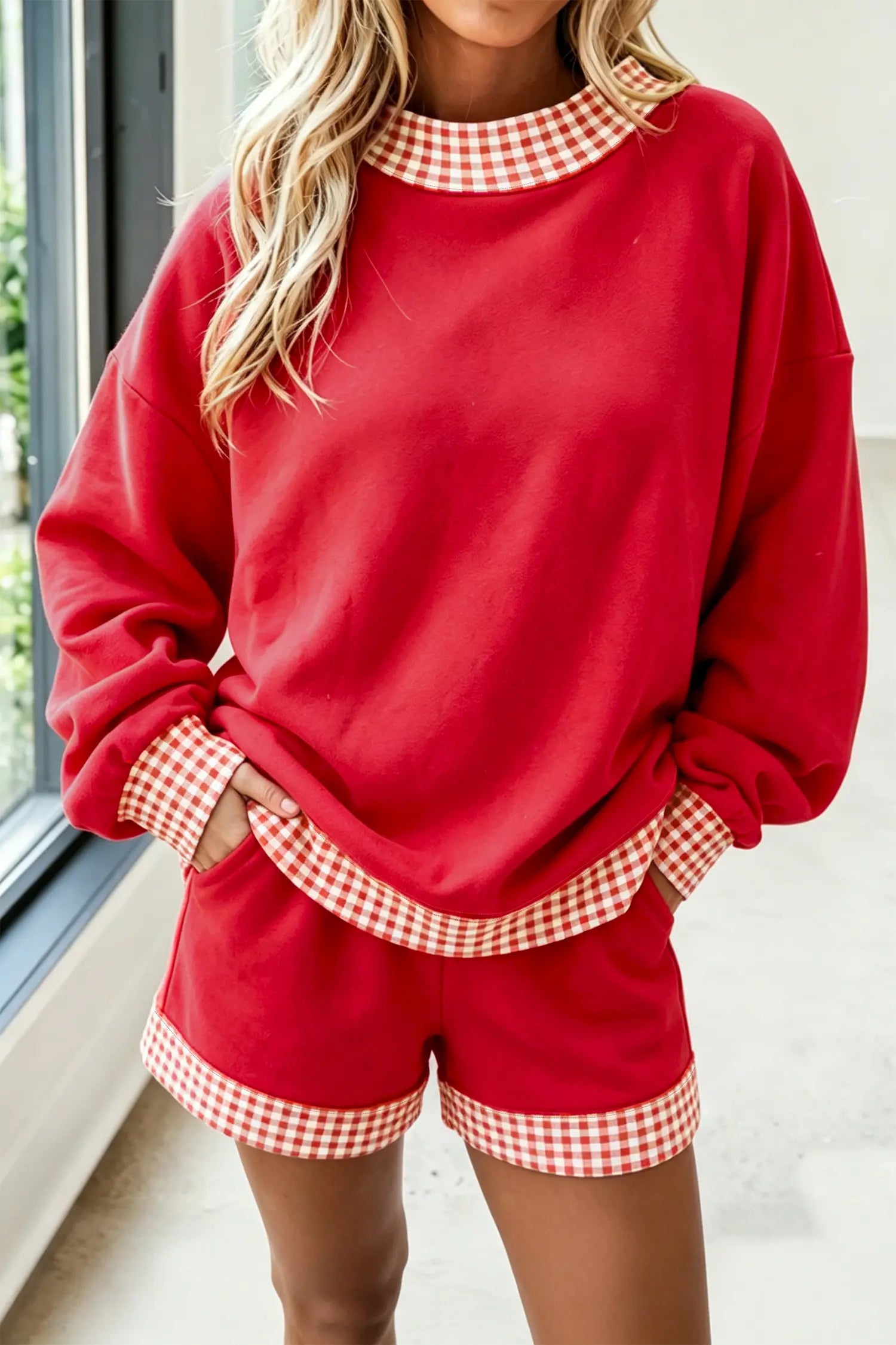 A woman wearing a red two-piece loungewear set with red and white gingham trim on the collar, cuffs, and shorts hems, standing near a window.