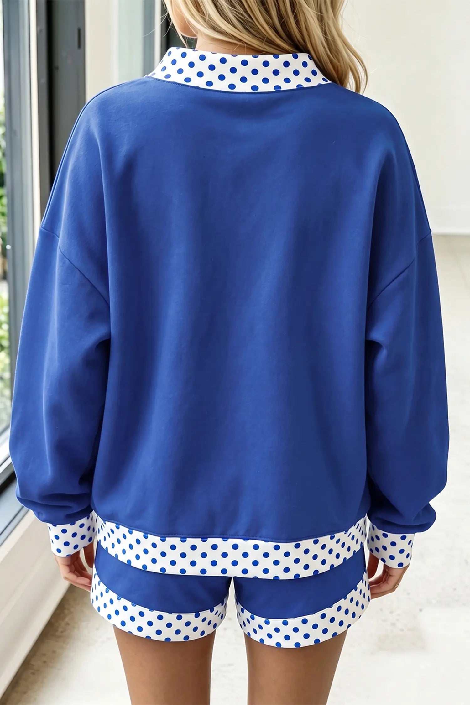 A person shown from the back wearing a royal blue sweatshirt and matching shorts set, both featuring white collar and hem details with blue polka dots.