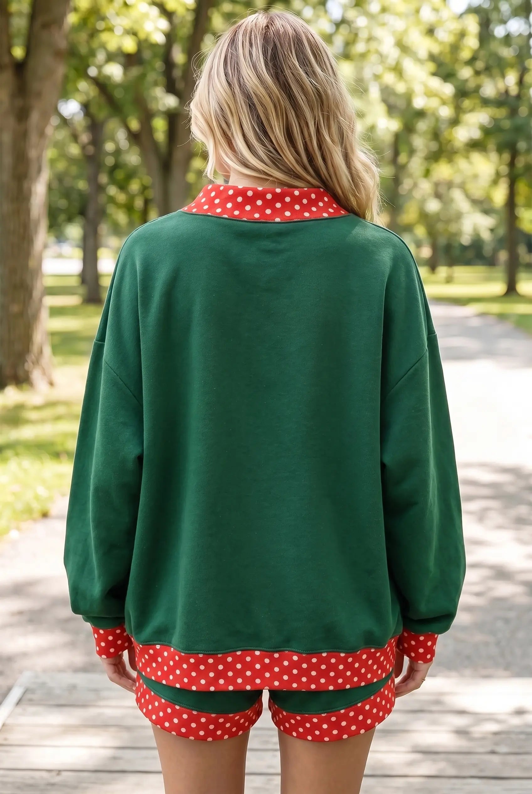 A woman viewed from behind wearing a dark green long-sleeve top and matching shorts with red and white polka dot trim, standing on a paved path in a park.
