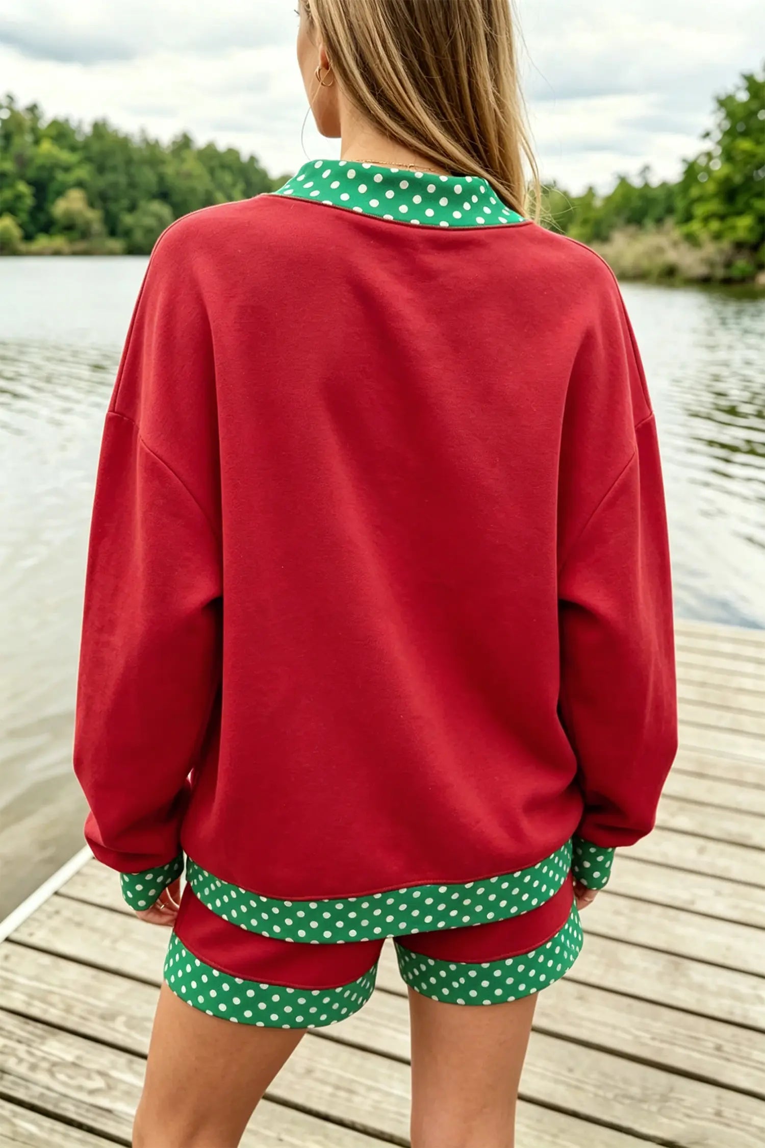 A woman viewed from behind wearing a red sweatshirt and matching shorts with green and white polka dot trim, standing on a wooden dock with water and trees in the background.