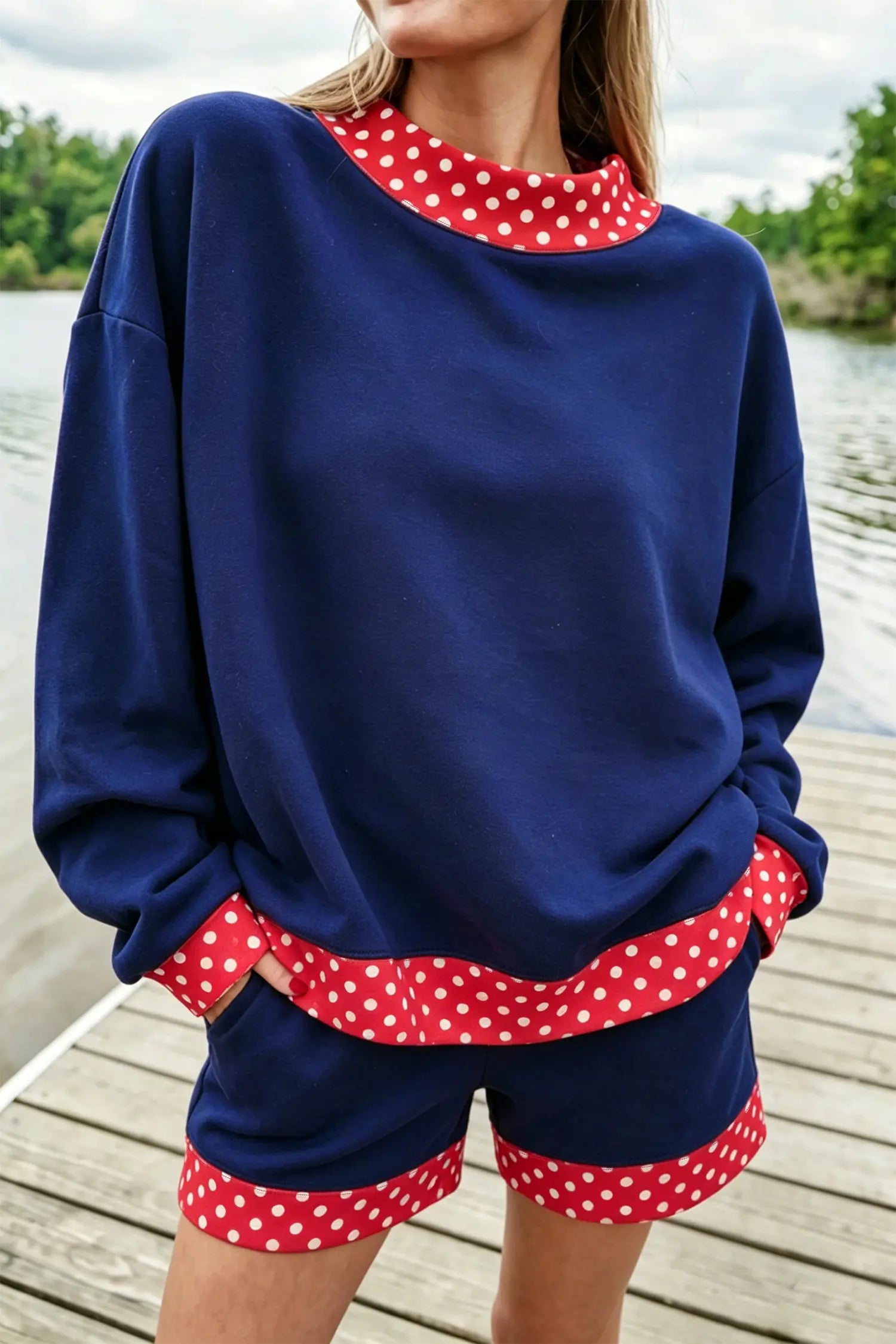 A woman wearing a navy blue lounge set with red and white polka dot trim, standing on a wooden dock with water and trees in the background.