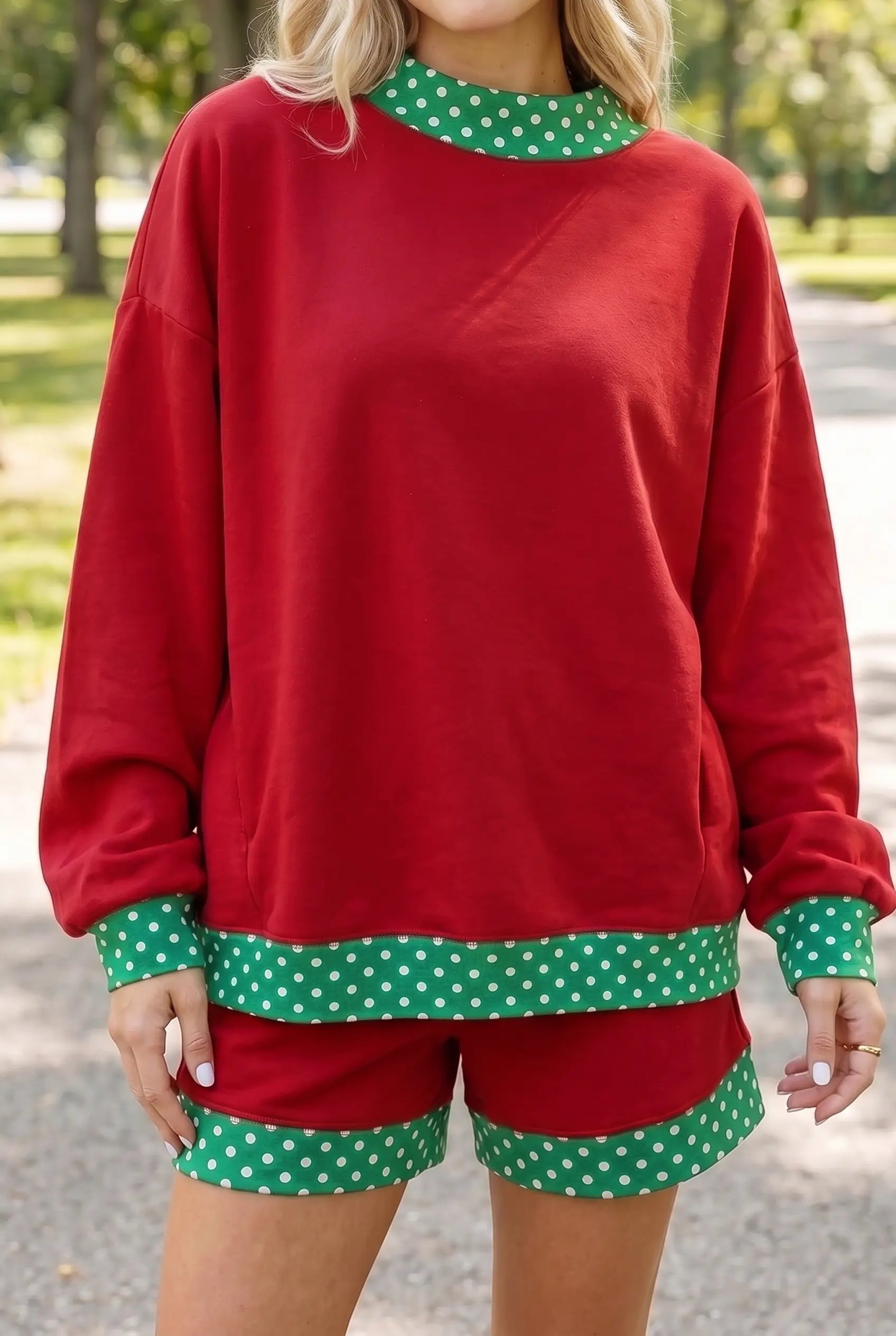 A woman wearing a red holiday-themed lounge set with green and white polka dot trim, featuring a long-sleeve sweatshirt and matching shorts, photographed outdoors on a paved path with trees in the background.
