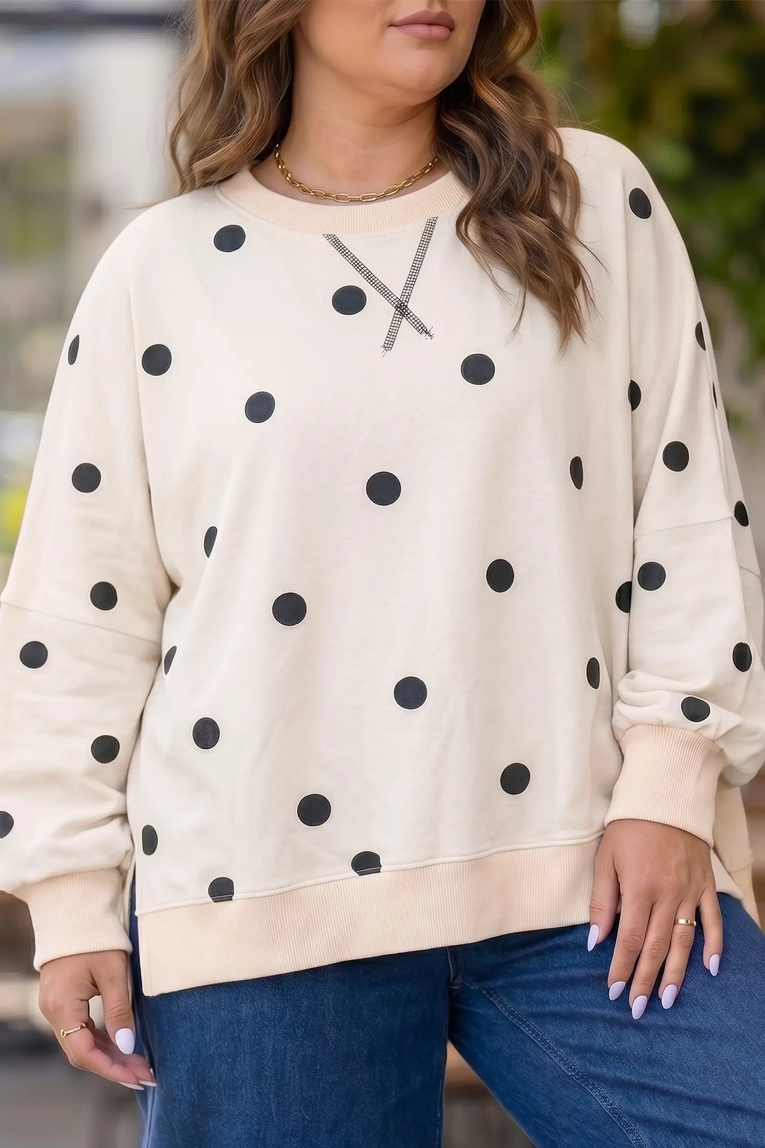 A woman wearing a cream-colored crewneck sweatshirt with black polka dots and a gray 'X' graphic on the chest, paired with blue jeans.