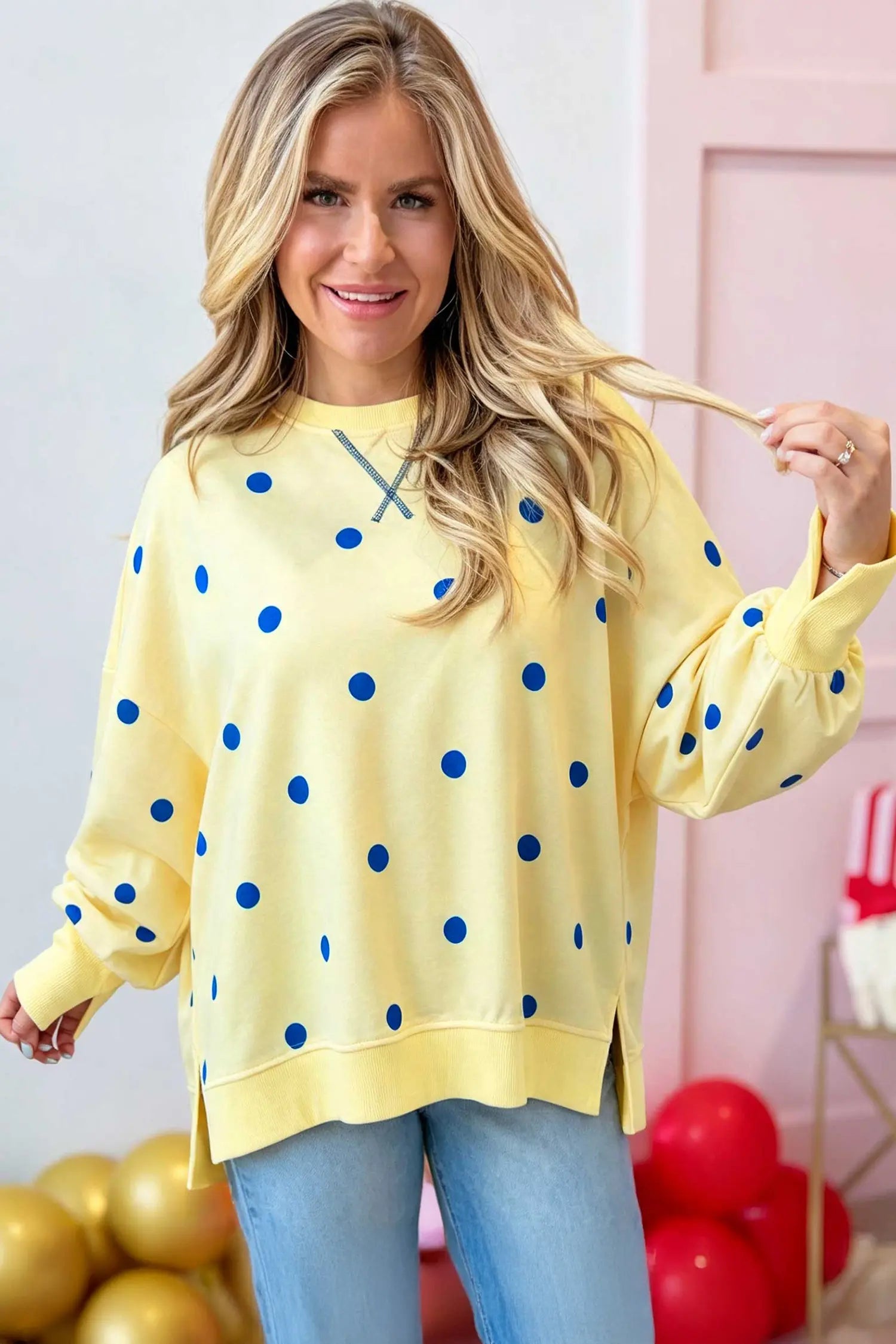 A woman wearing a pale yellow long-sleeve sweatshirt with blue polka dots, paired with light blue jeans, standing in a decorated room with balloons.