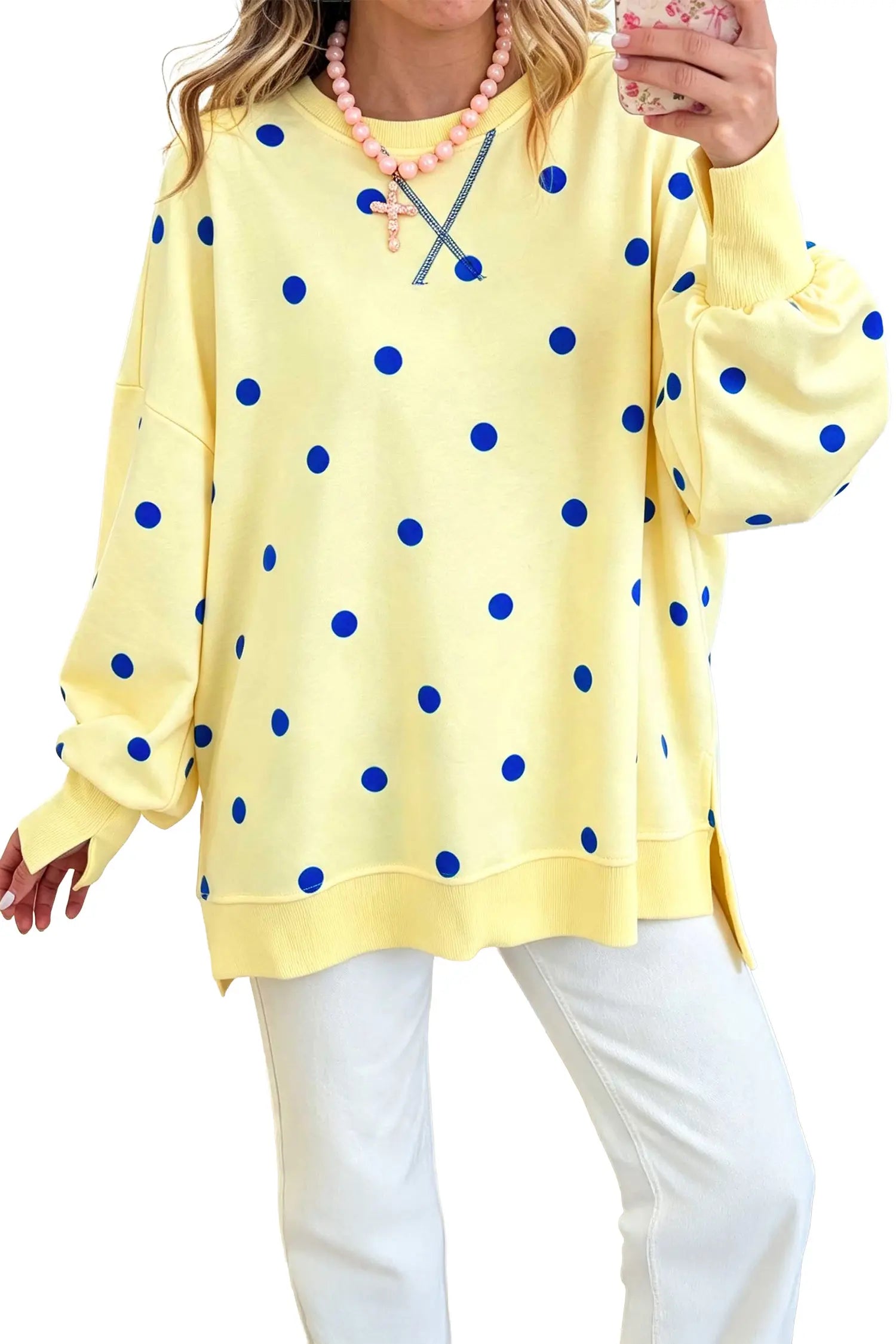 A woman wearing a pale yellow long-sleeve sweatshirt with blue polka dots, paired with white pants and a pink beaded necklace, holding a phone in a mirror selfie.