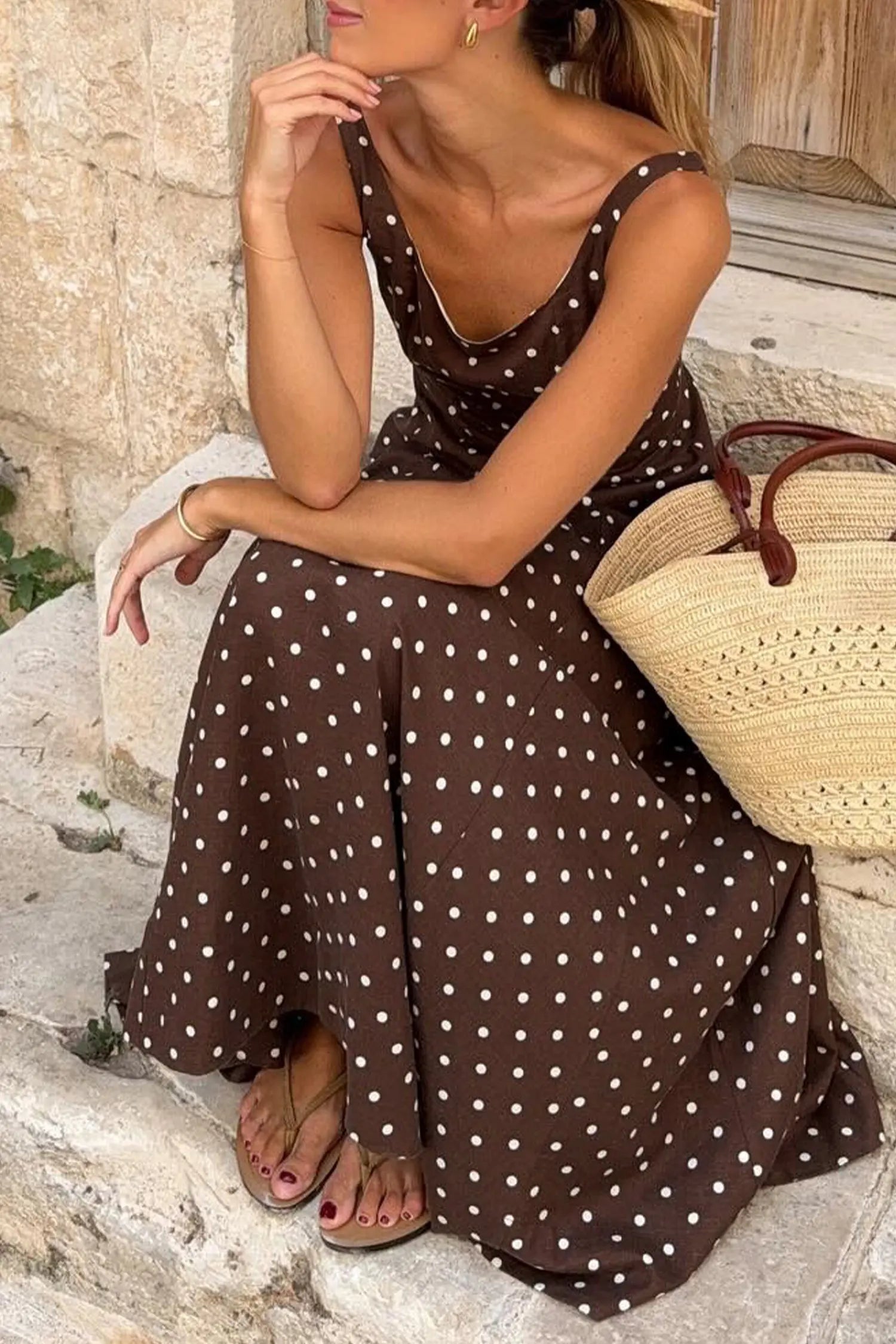 Woman in a brown polka dot dress sitting on stone steps with a straw bag.