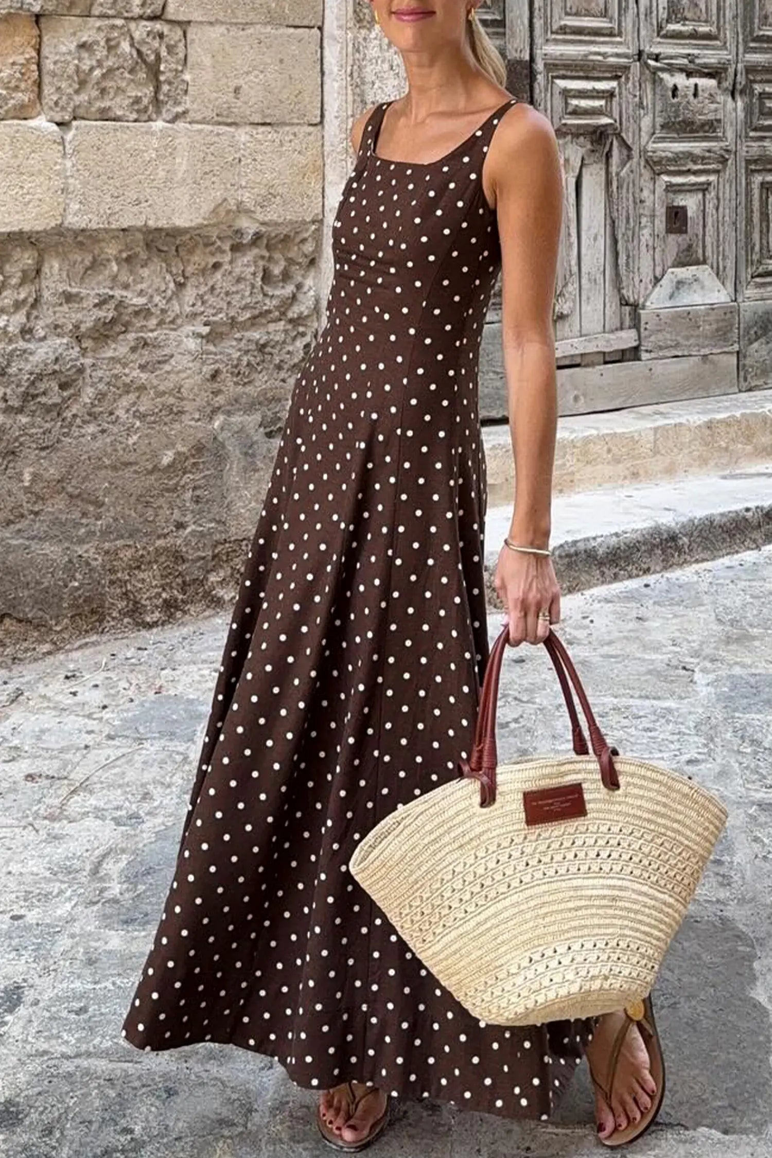 Woman in a brown polka dot dress holding a straw bag against a stone wall.