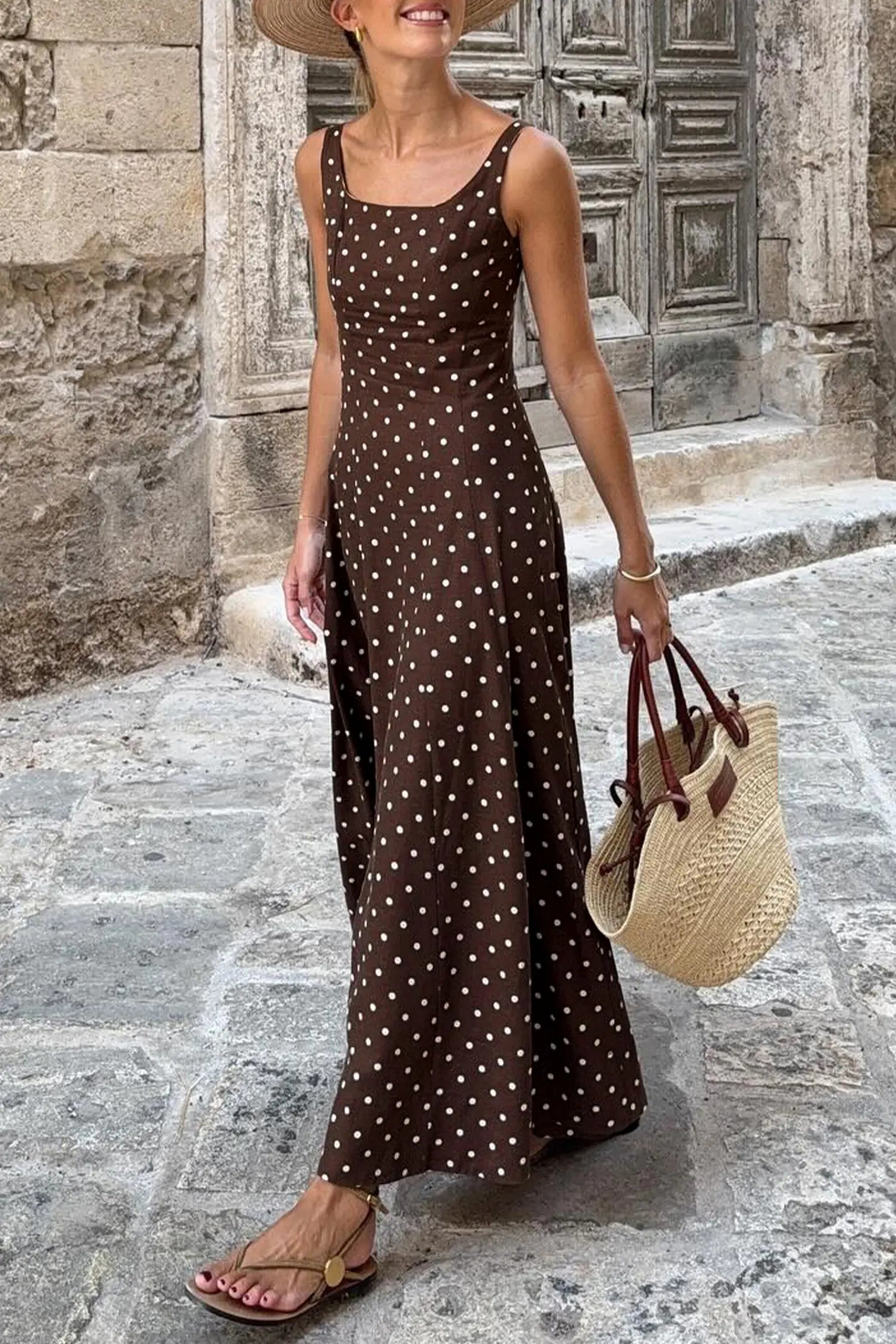 Woman in a brown polka dot dress holding a straw bag on a stone street.