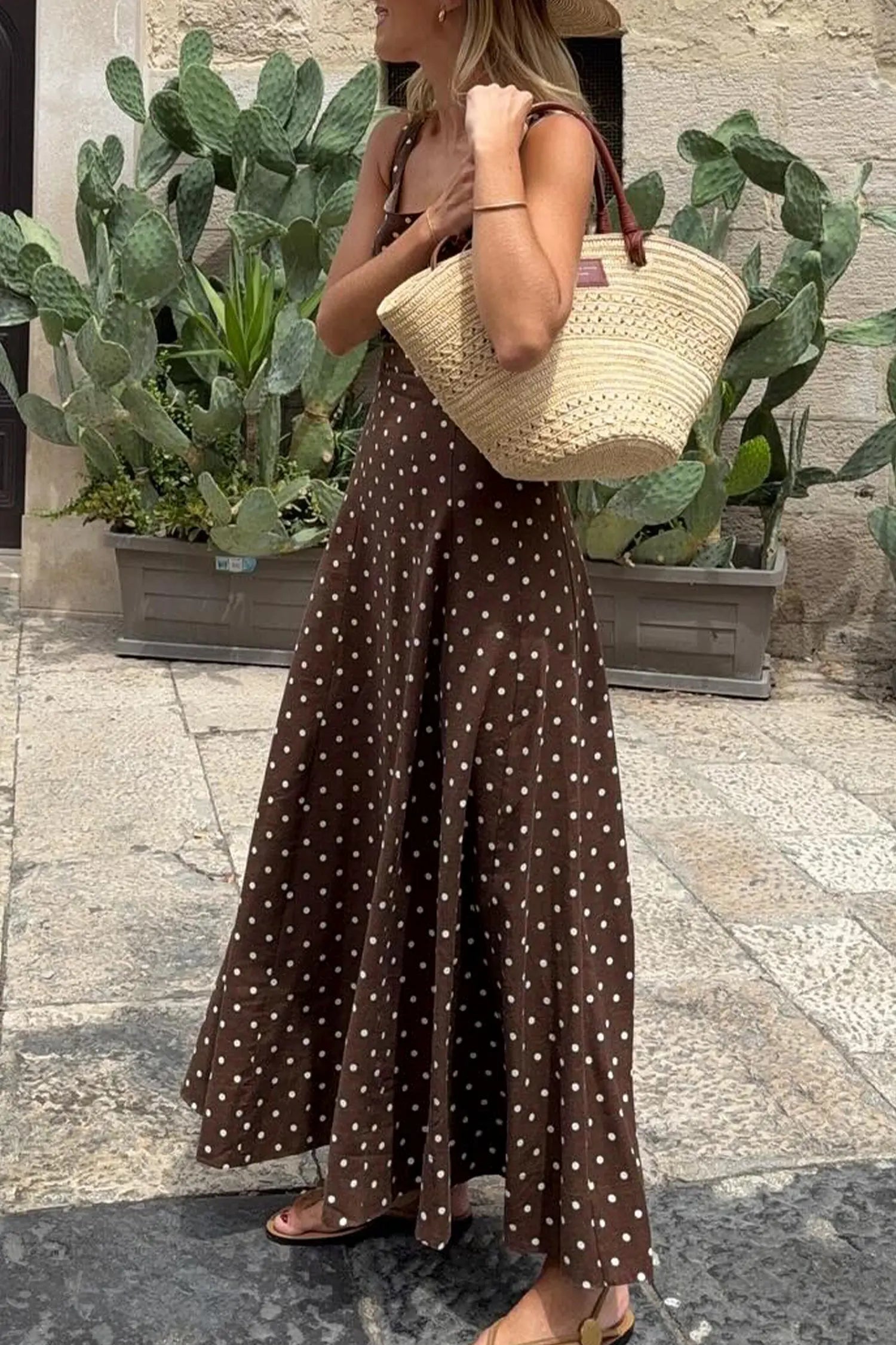 Woman in a brown polka dot dress holding a straw bag with plants in the background