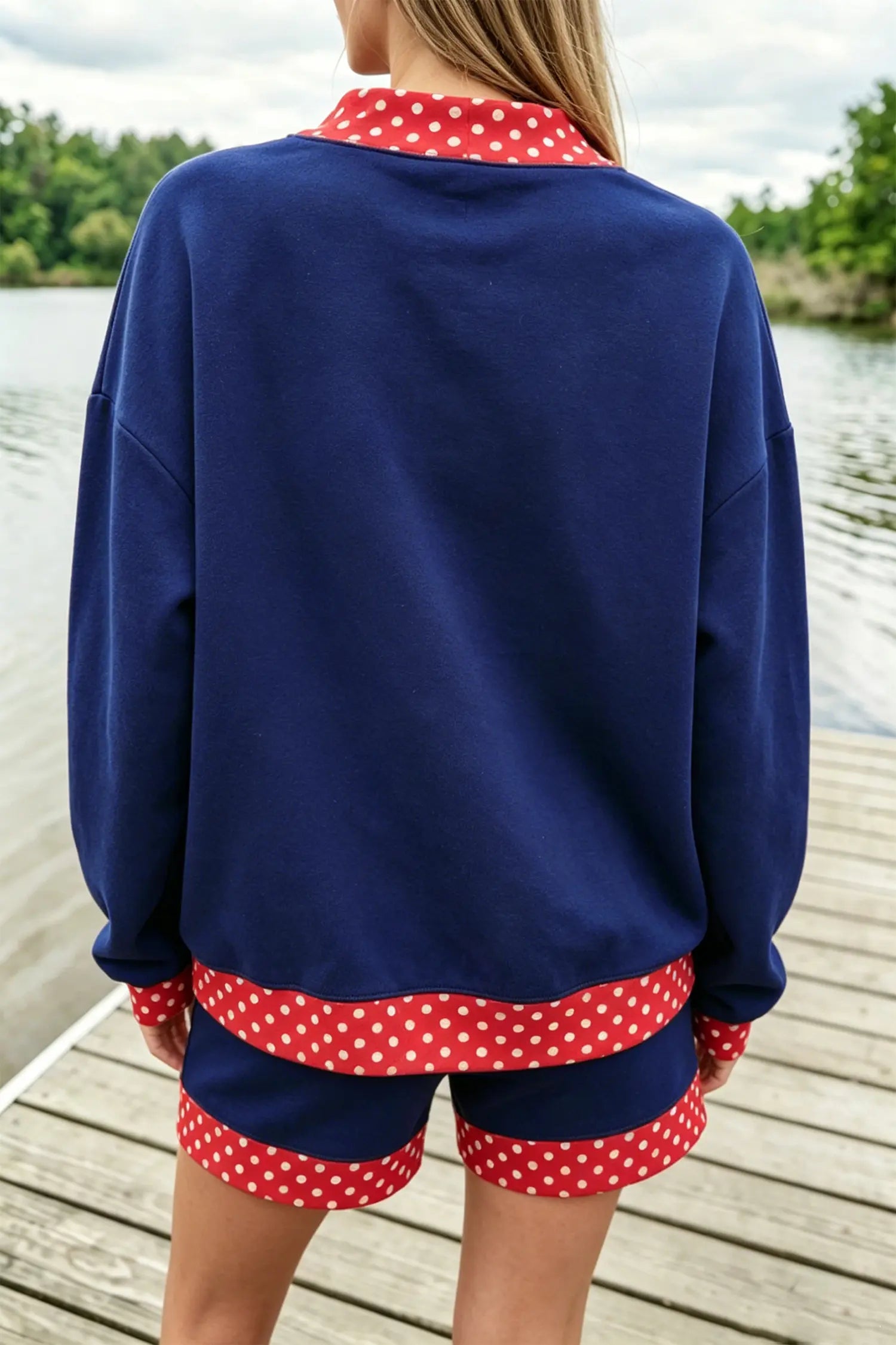 A person viewed from behind wearing a navy blue lounge set with red and white polka dot trim, standing on a wooden dock with water and trees in the background.