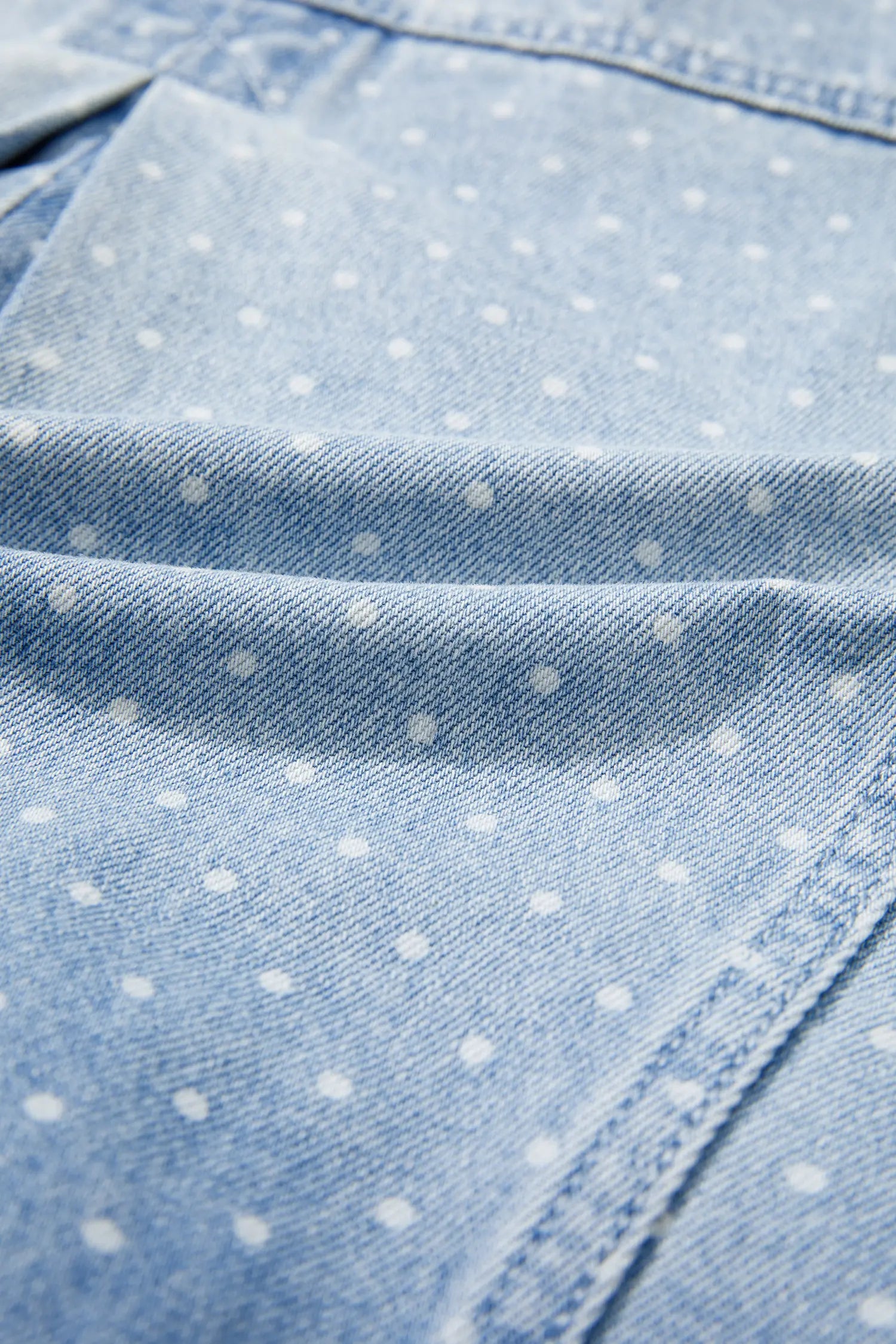 Close-up of light blue denim fabric with a white polka dot pattern and visible stitching.