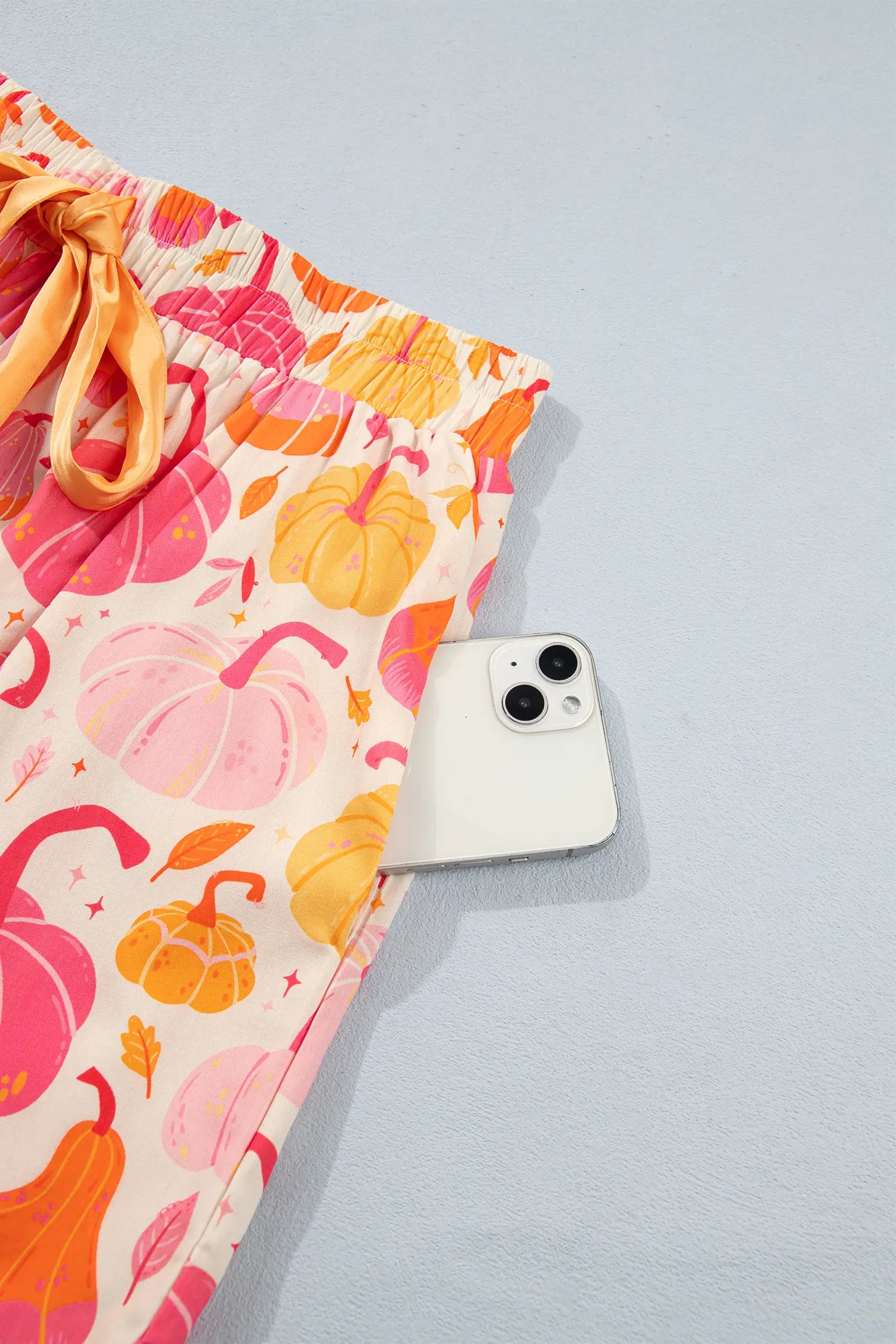 A close-up of a pair of shorts with a colorful autumn pumpkin print, featuring orange and pink pumpkins on a cream background with a matching orange drawstring. A white smartphone is placed next to the shorts for scale, set against a light blue-gray background.