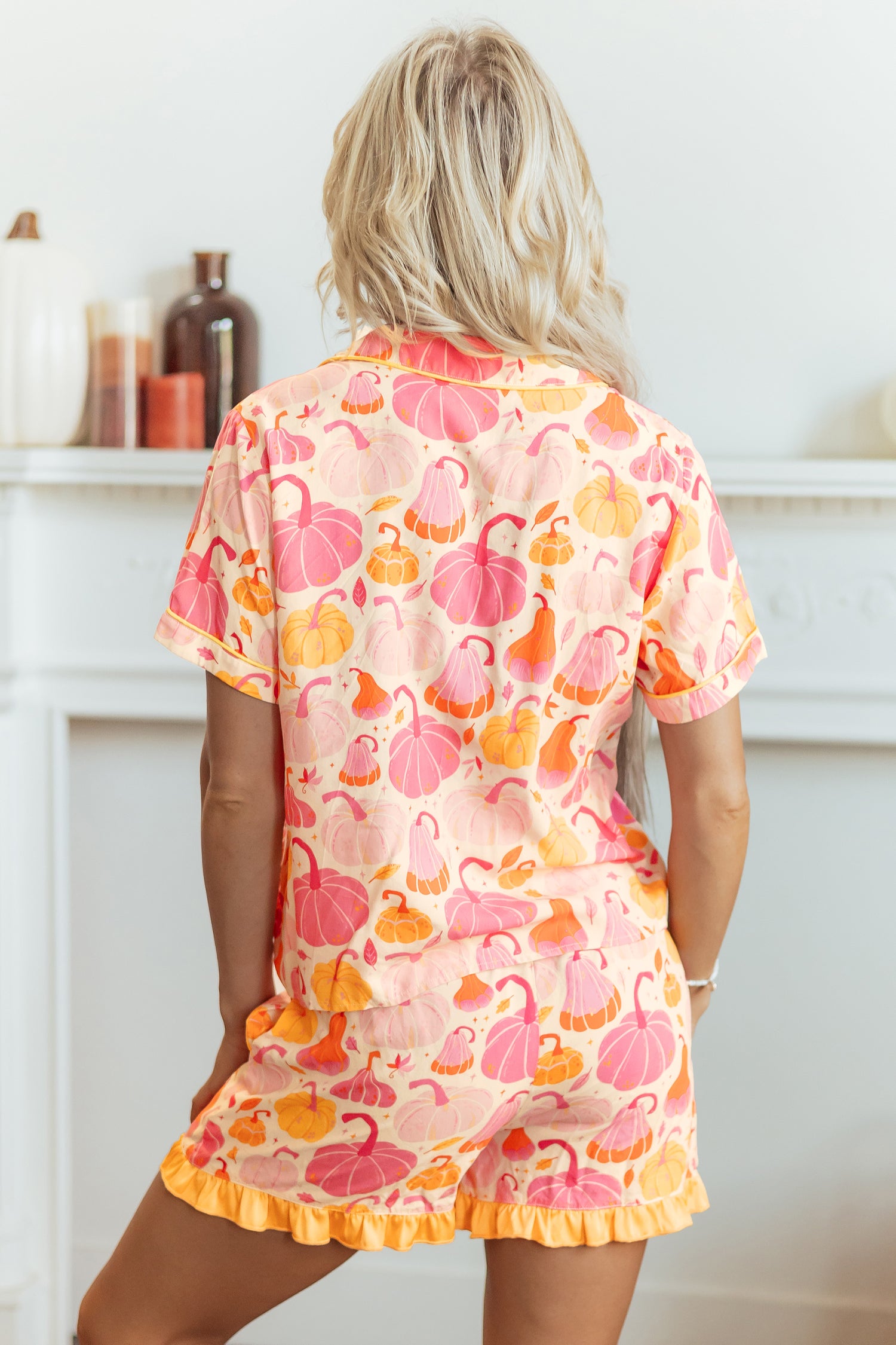 A woman viewed from behind wearing a short-sleeve ruffled romper with a pink, orange, and yellow pumpkin pattern on a light peach background, posed in front of a white fireplace mantel.