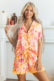 A woman modeling a short-sleeve pajama romper with a pink, orange, and yellow flamingo print, standing in a brightly lit room with a white fireplace background.