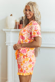 A woman modeling a short-sleeve pajama robe and matching shorts set with a colorful fall harvest print of pumpkins, gourds, and acorns in orange, pink, and yellow on a light peach background, set against a white fireplace background.