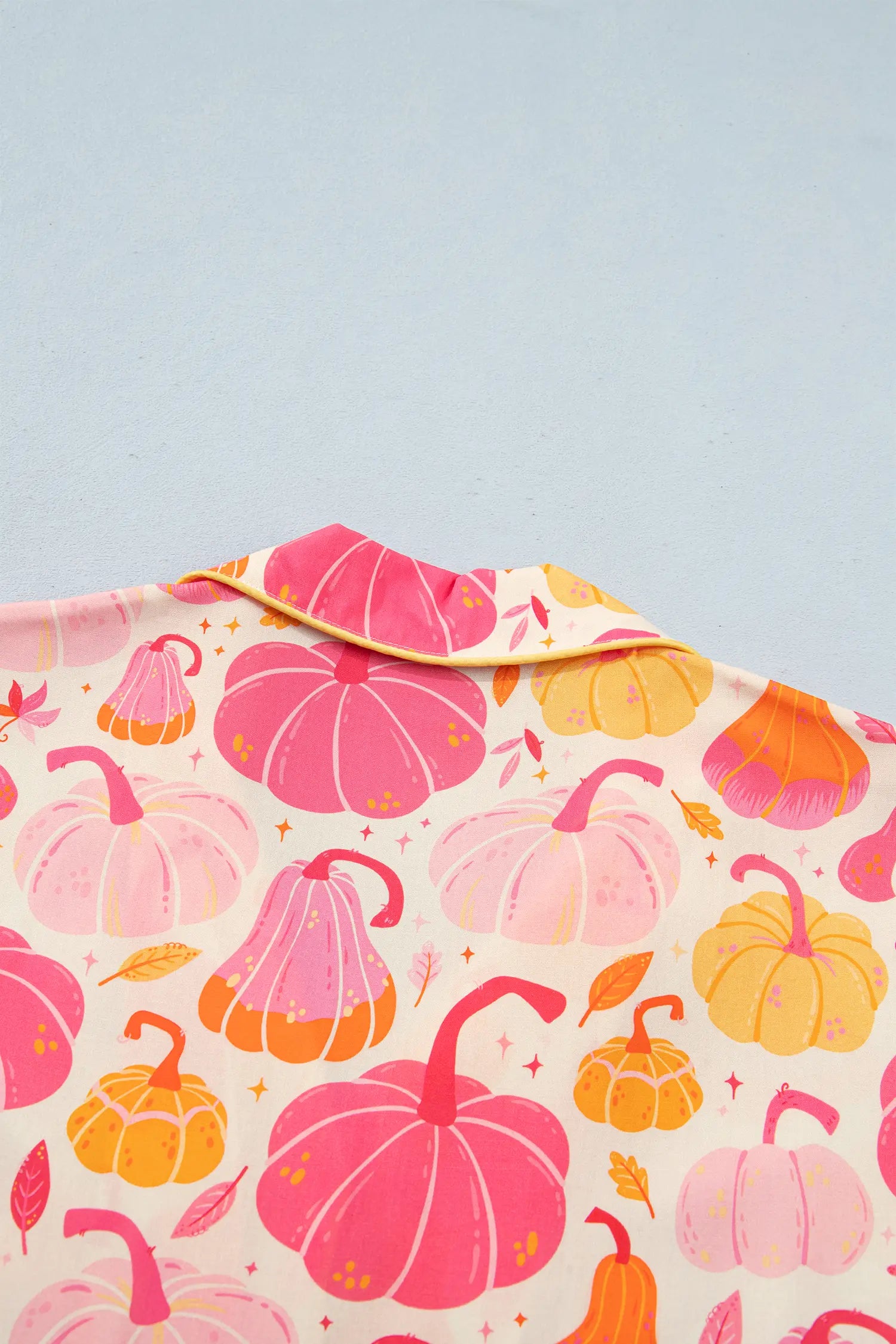 A close-up of a fabric product, likely a shirt collar or garment panel, featuring a colorful autumn pumpkin print on a cream background. The print includes various shades of pink, orange, and yellow pumpkins with small gold stars and leaves, set against a solid light blue-gray background.