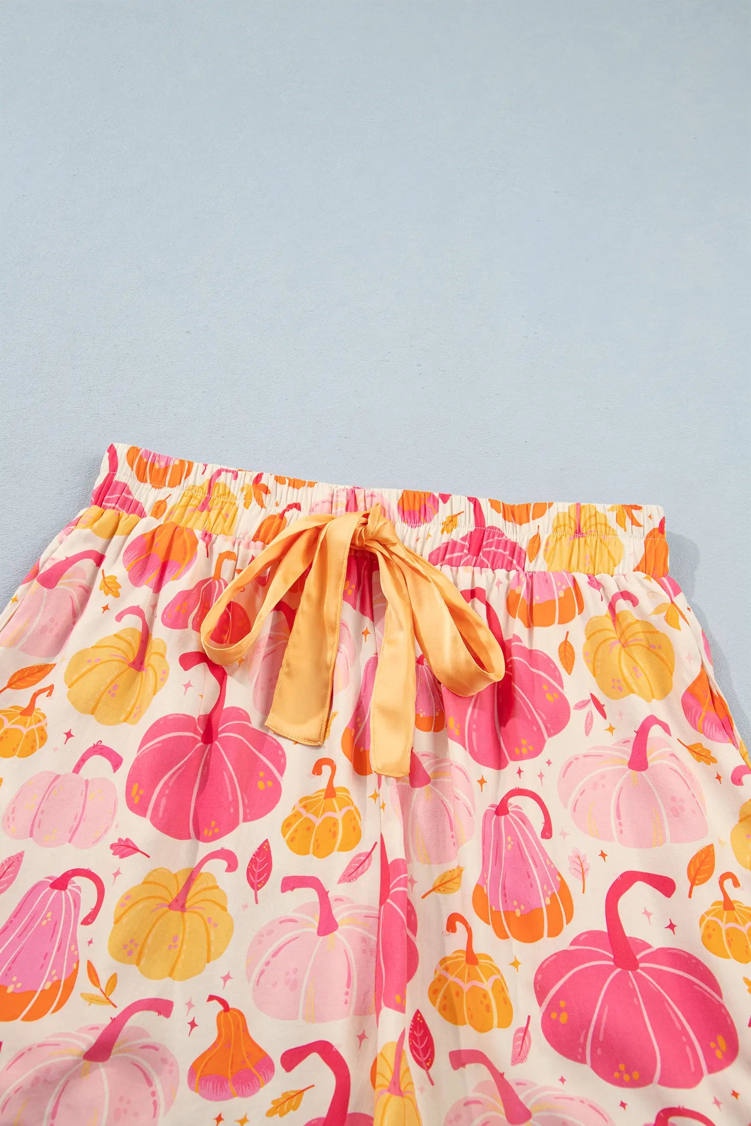 A close-up of a pair of shorts with an elastic waistband and a matching orange drawstring, featuring a colorful print of pumpkins in pink, orange, and yellow on a cream background.