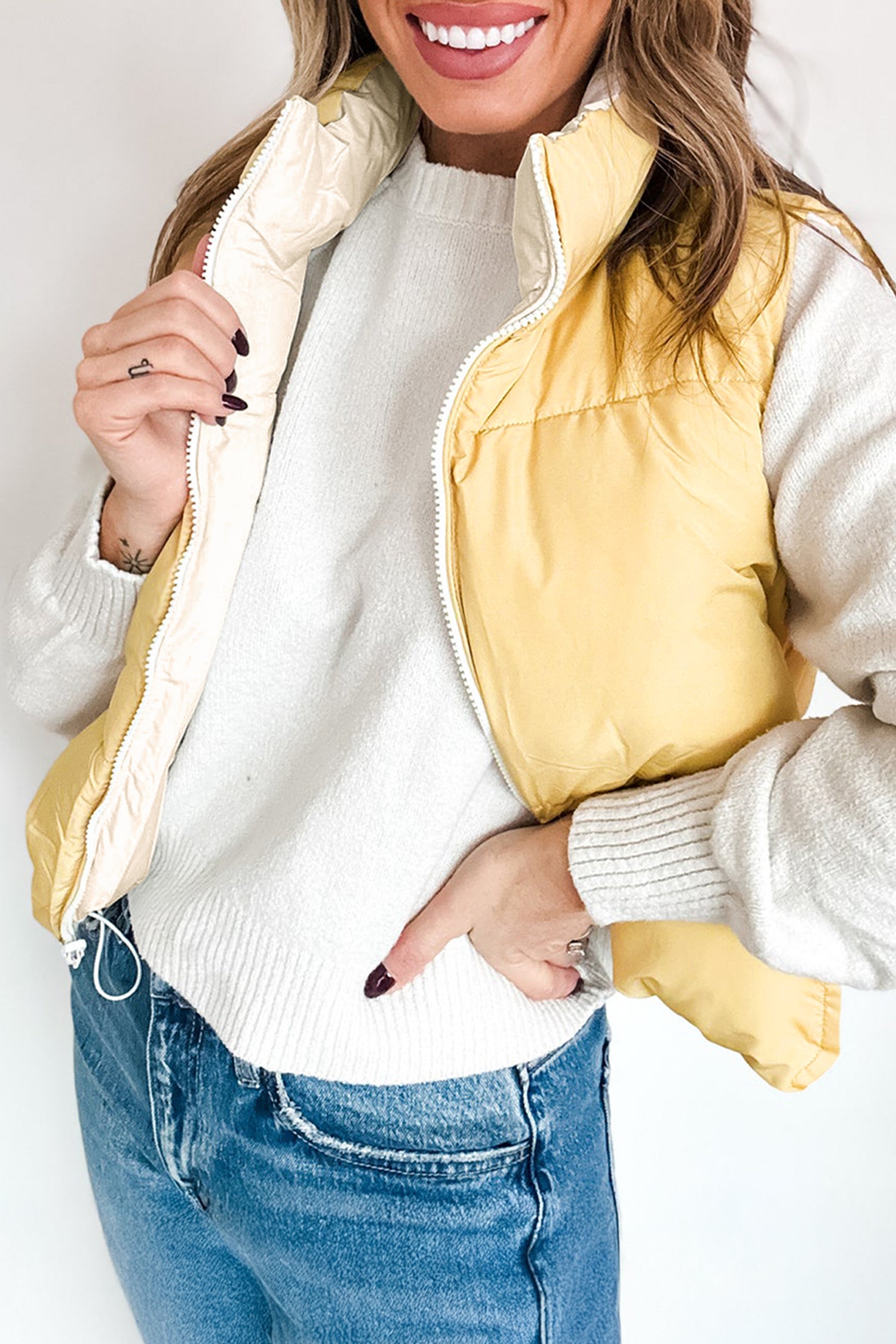 Woman in pale yellow puffer vest over white knit sweater and blue jeans against a light background.