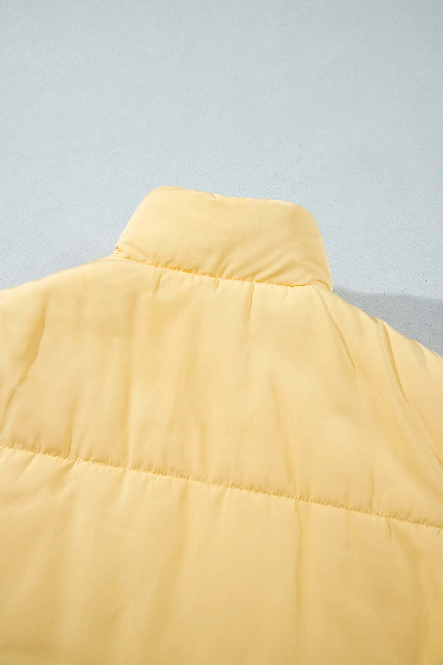Close-up of pale yellow puffer jacket with padded collar and horizontal quilting on light gray background.