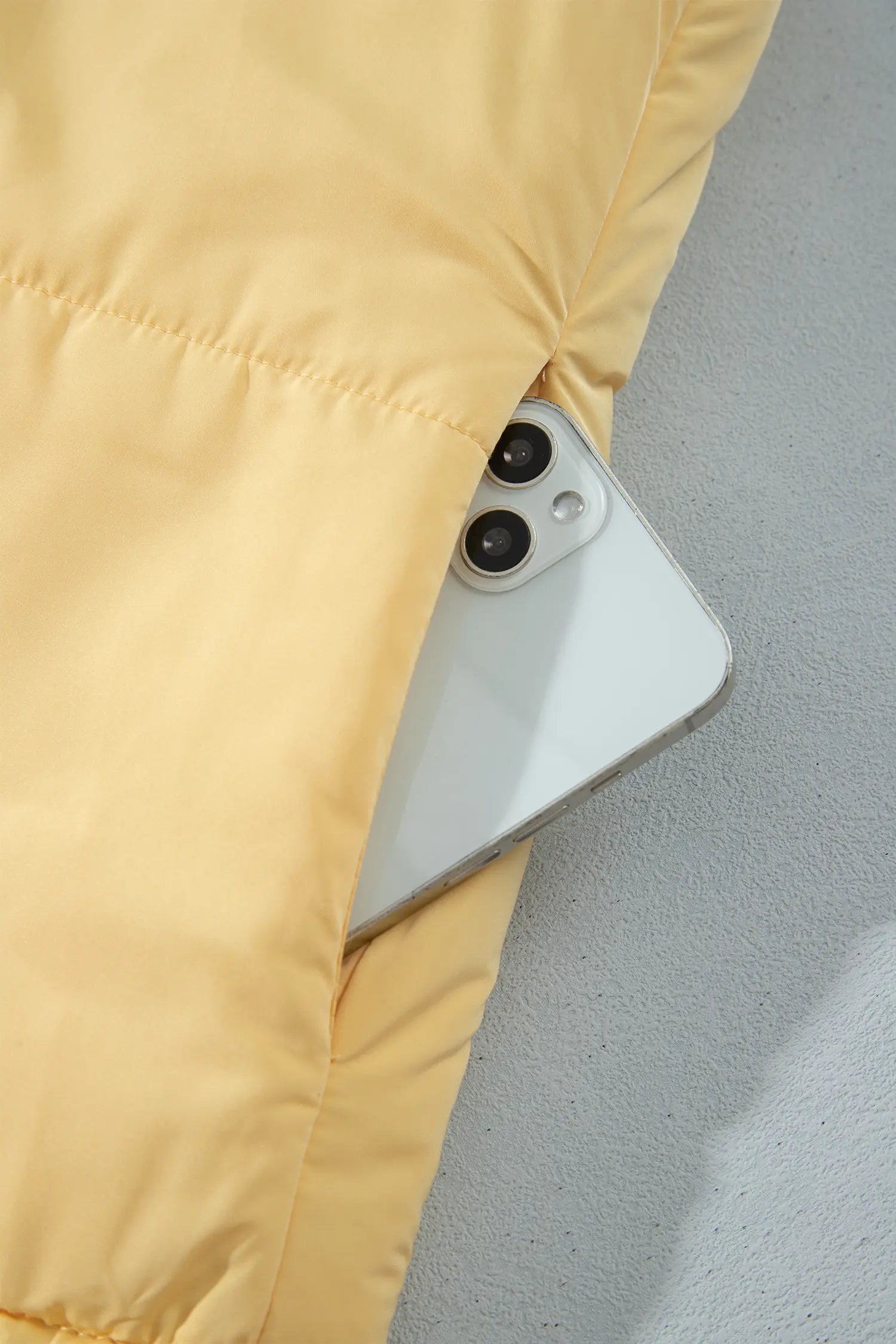 Close-up of light yellow puffer jacket sleeve with a white smartphone tucked inside against gray.