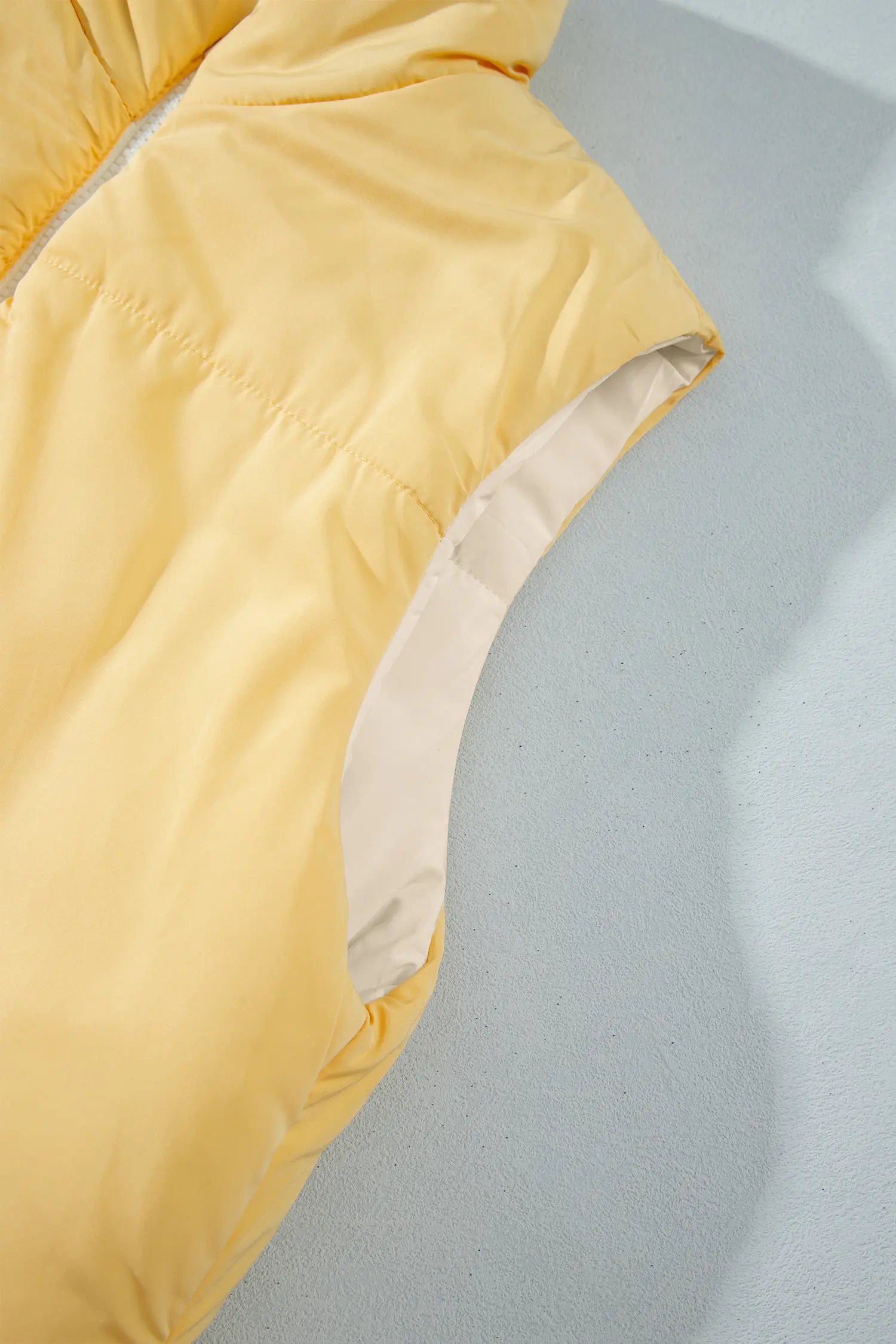 Close-up of light yellow puffer vest with smooth shell and contrasting beige lining on gray surface.