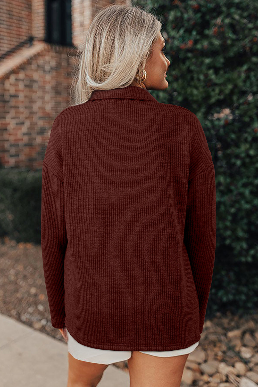 Woman wearing a brown textured sweater in front of a brick wall and green bush