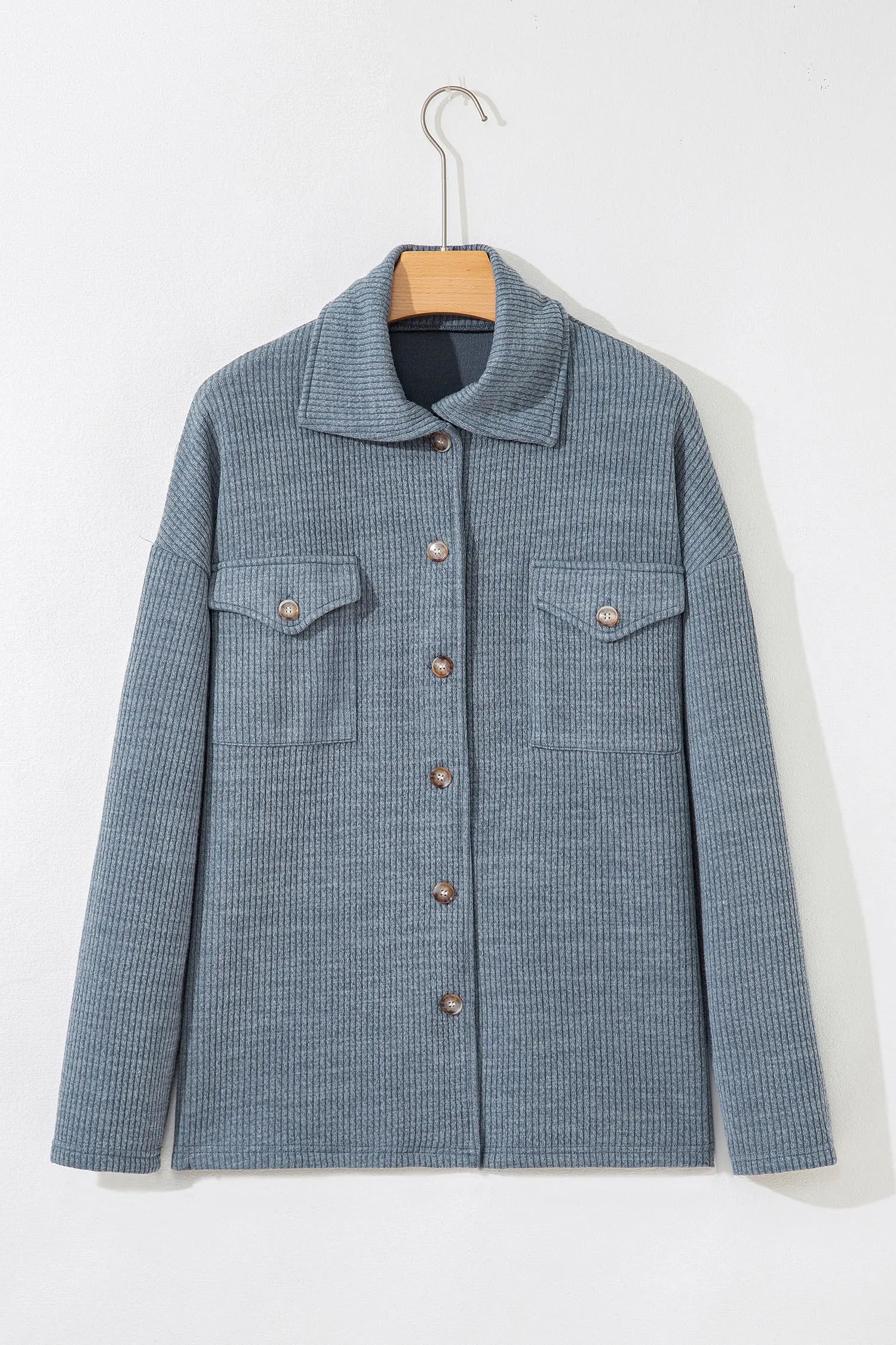 A textured blue-gray button-up jacket with a pointed collar and two chest flap pockets, hanging on a wooden hanger against a plain white wall.