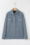 A textured blue-gray button-up jacket with a pointed collar and two chest flap pockets, hanging on a wooden hanger against a plain white wall.