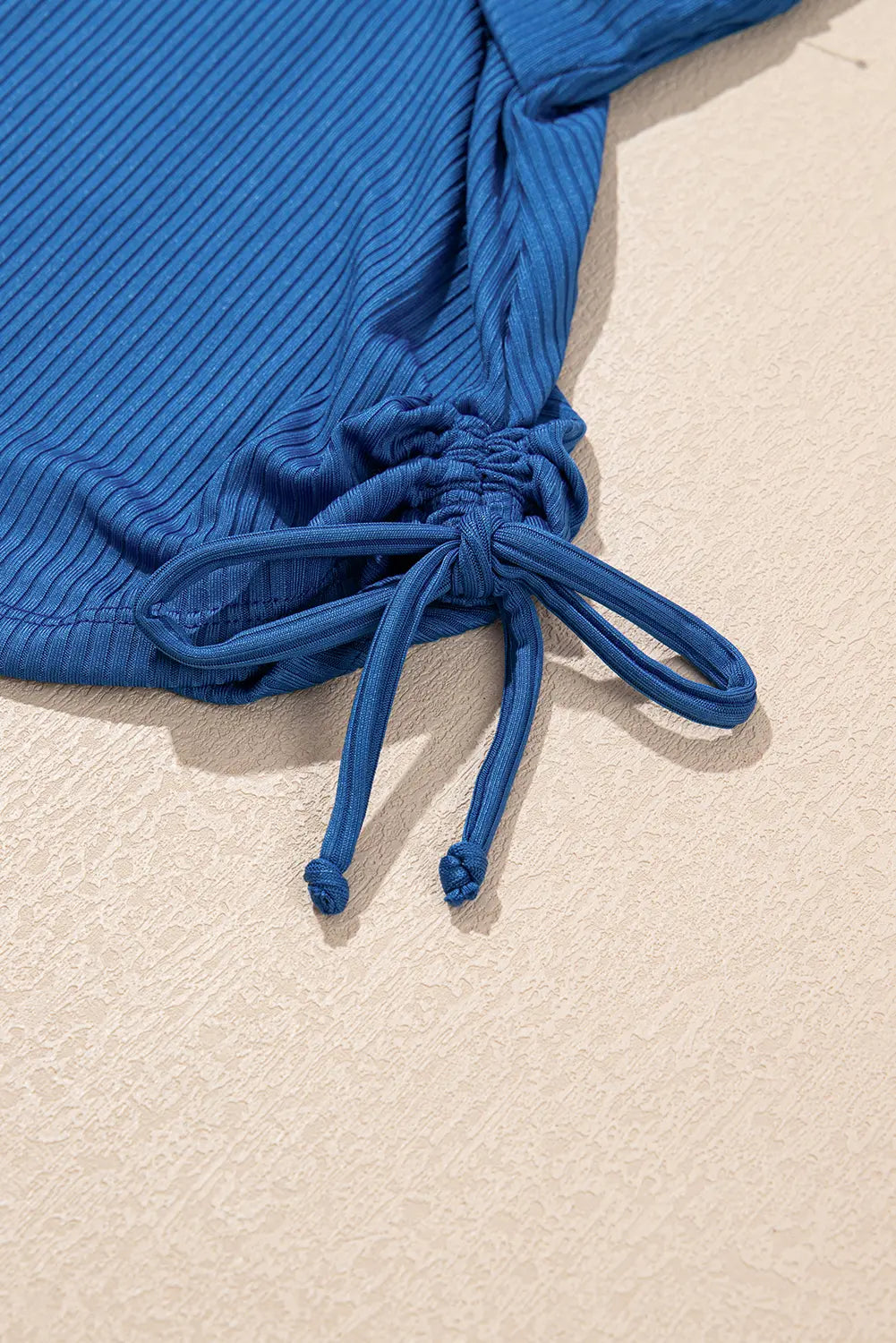 Blue ribbed swimsuit top with drawstring on a beige surface