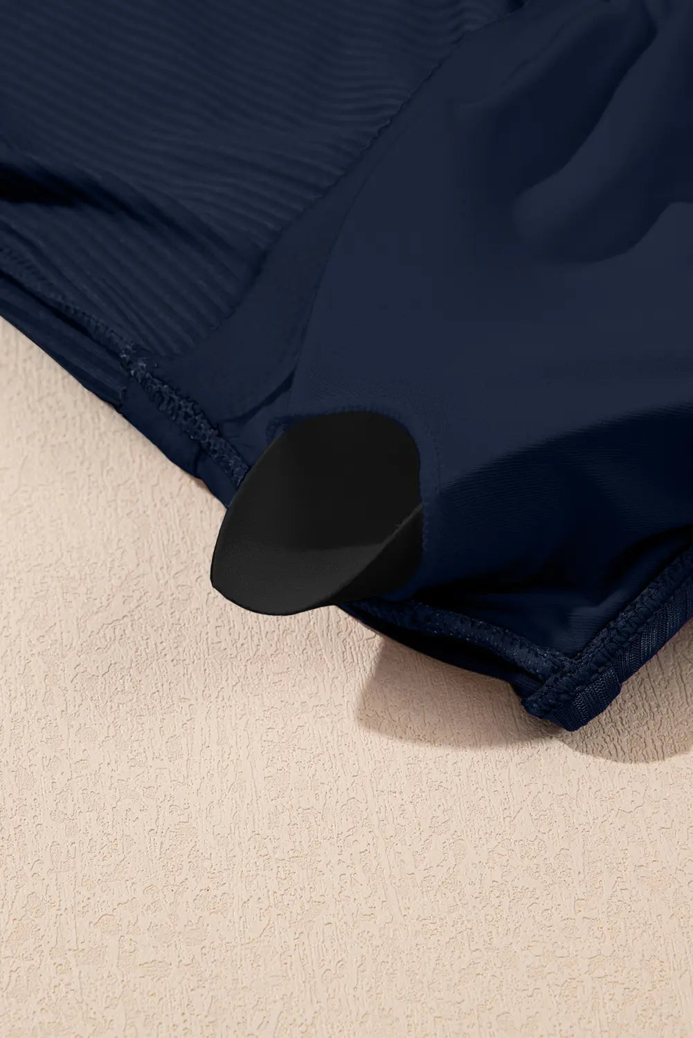 Close-up of a navy blue ribbed fabric garment laid on a textured beige surface, with a black curved trim or accent visible near the lower edge.
