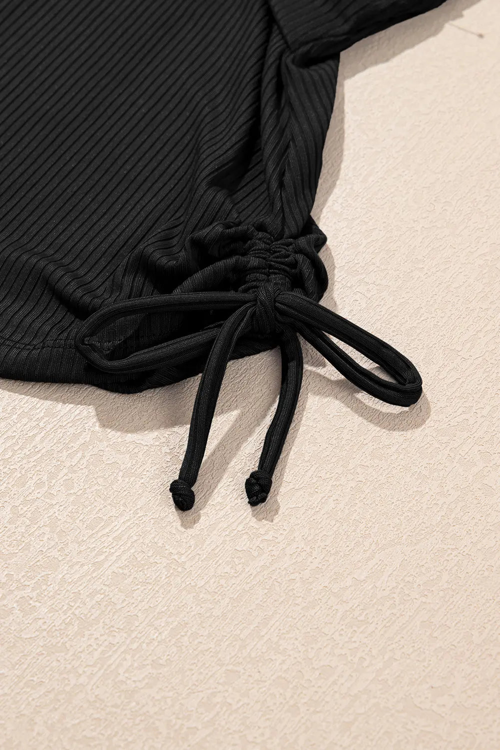 Close-up of a black ribbed drawstring garment laid on a light beige textured surface, showing the fabric texture and the tie detail.