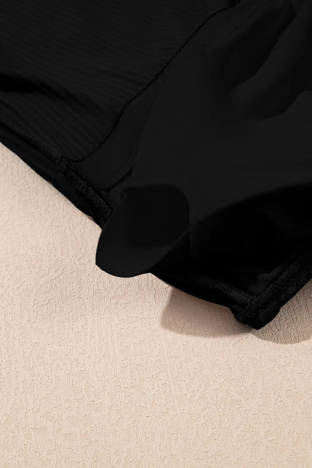 Close-up of a black garment fabric with a ribbed texture and a draped hem, resting on a textured beige surface.