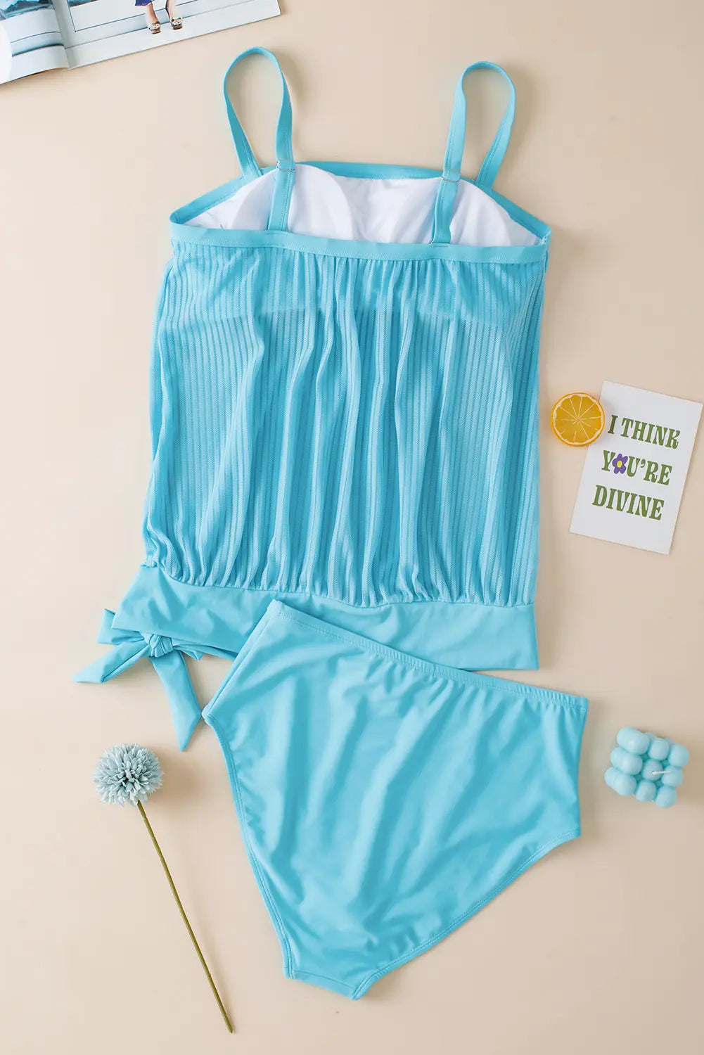A two-piece baby girl's swimsuit in light blue, featuring a sleeveless top with thin shoulder straps, a gathered pleated front, and a side tie, paired with matching high-cut bottoms. The swimsuit is laid flat on a beige background with decorative props including a dandelion, a small card, and a lemon slice.