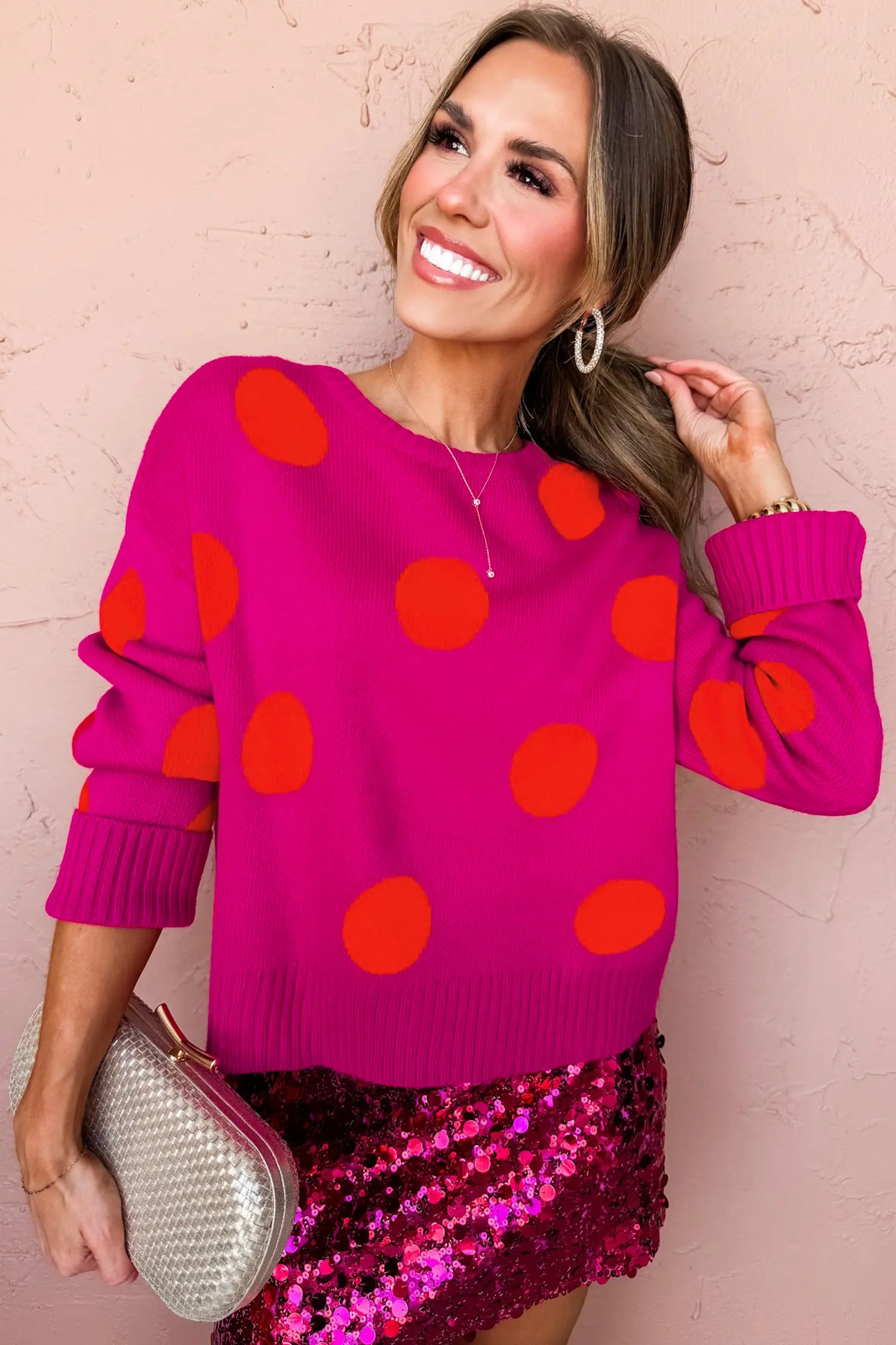 A woman wearing a bright pink crewneck sweater with large orange-red polka dots, paired with a shiny magenta sequined mini skirt and holding a silver clutch, posing against a textured pink wall.
