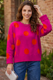 A woman wearing a bright pink knit sweater with large orange polka dots, paired with blue jeans, holding a silver clutch in front of a brick wall background.