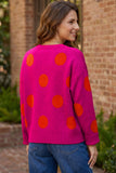 A woman wearing a bright pink knit sweater with large orange polka dots, shown from the back in an outdoor setting with a brick wall background.