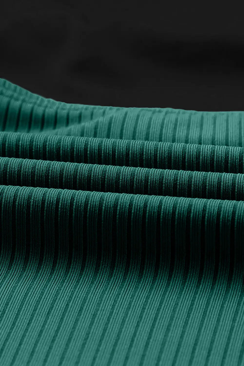A close-up of dark teal green ribbed fabric with horizontal folds, set against a solid black background.