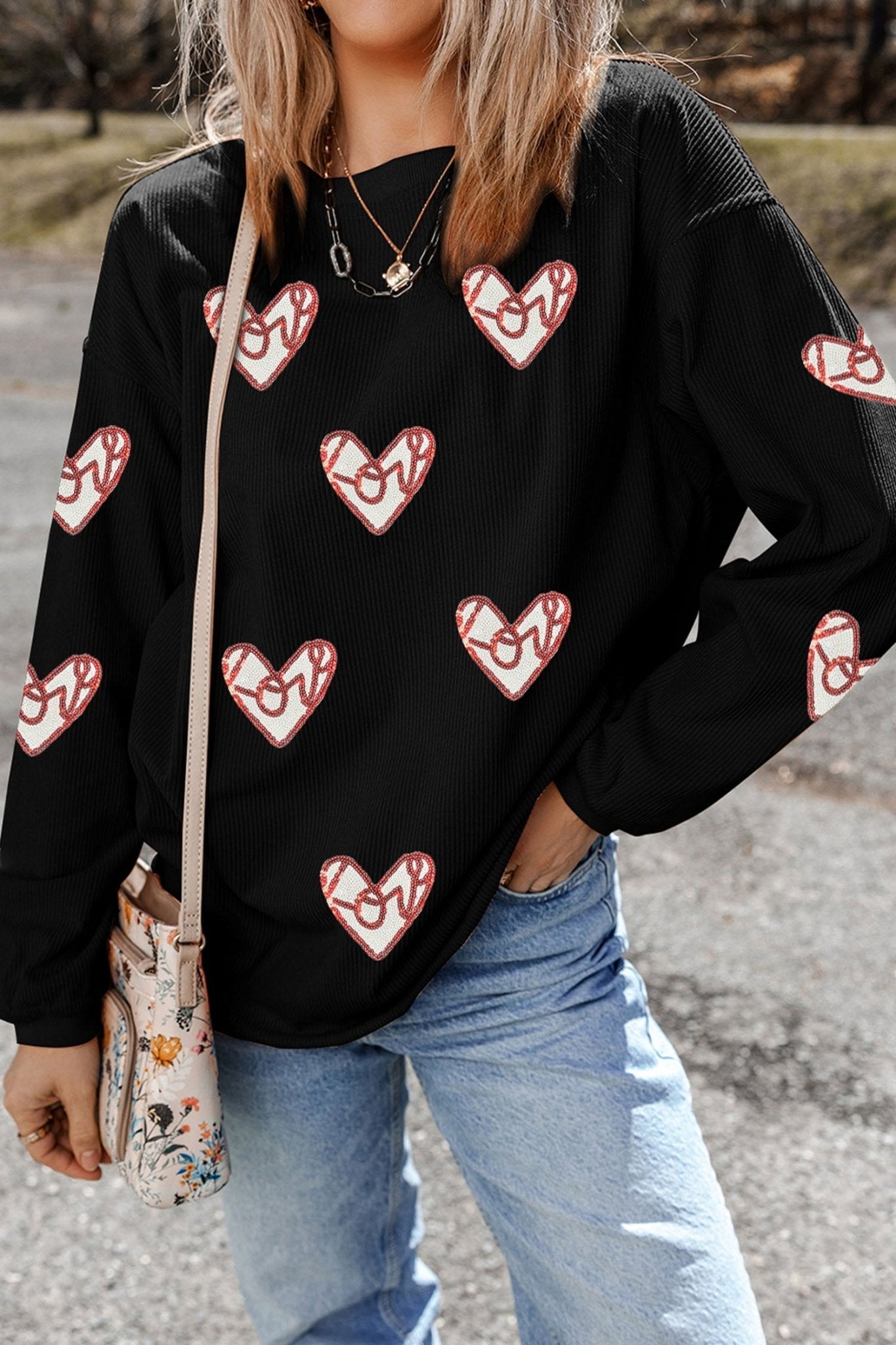 Person wearing a black sweatshirt with heart patterns, holding a floral bag outdoors.