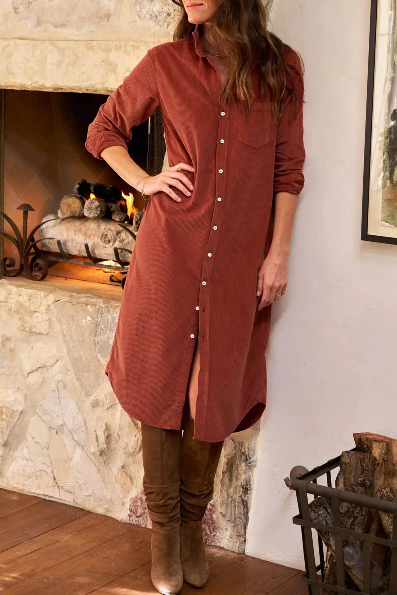 A woman wearing a rust-colored corduroy shirt dress with brown suede boots, standing in front of a stone fireplace.