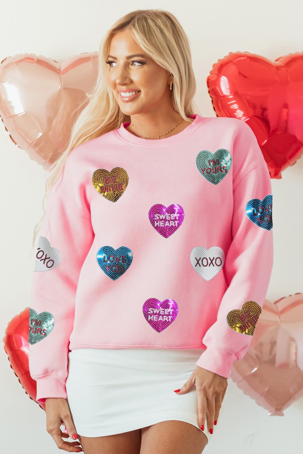 Woman wearing a pink sweatshirt with heart-shaped decorations in front of heart balloons.
