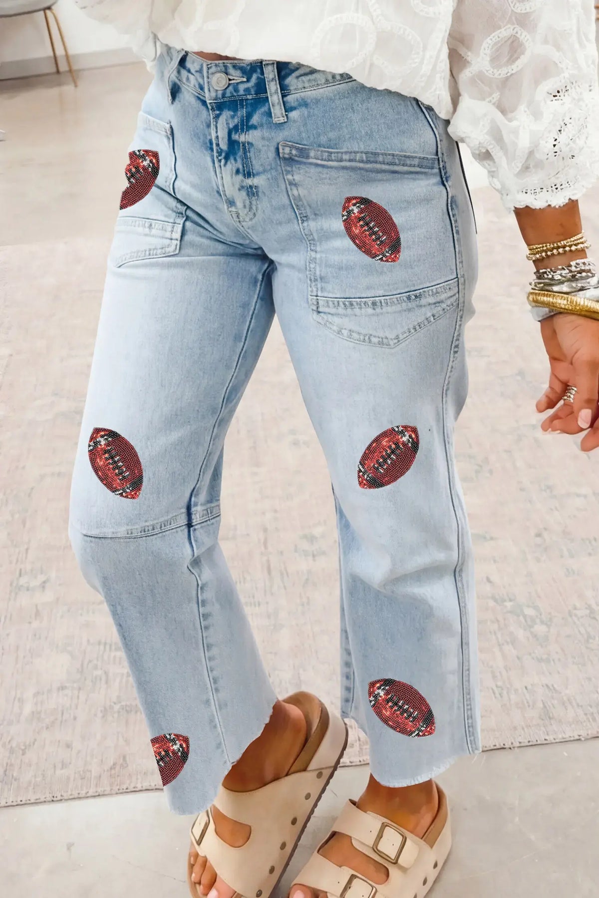 Light wash blue denim jeans with embroidered football patches on the thighs and lower legs, shown on a model wearing a white lace-trim top and beige slide sandals.